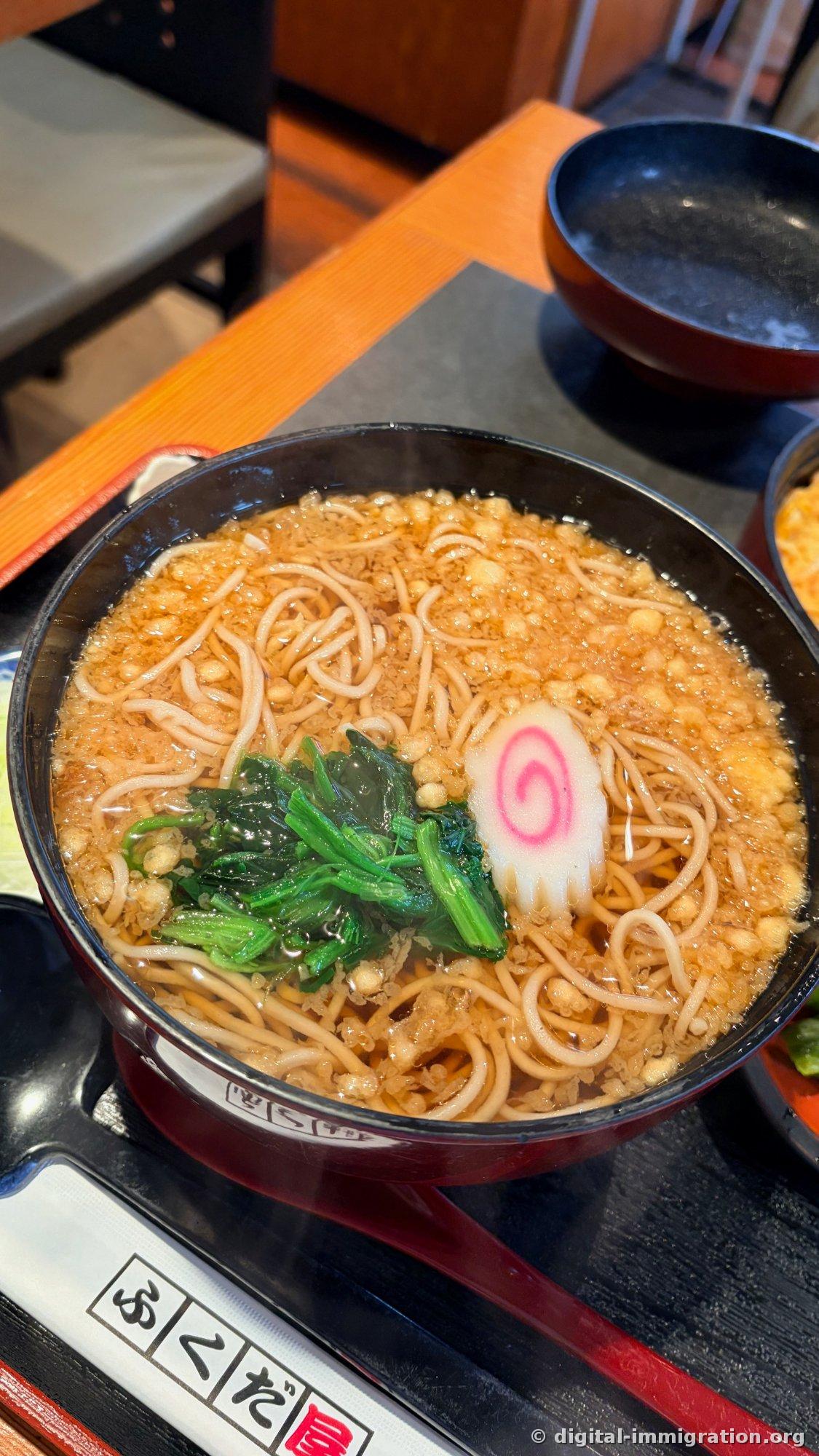 Soba chaud dans un bol noir. Les miettes de tempura couvrent le bouillon avec des épinards blanchis et du surimi rose tourbillon dessus