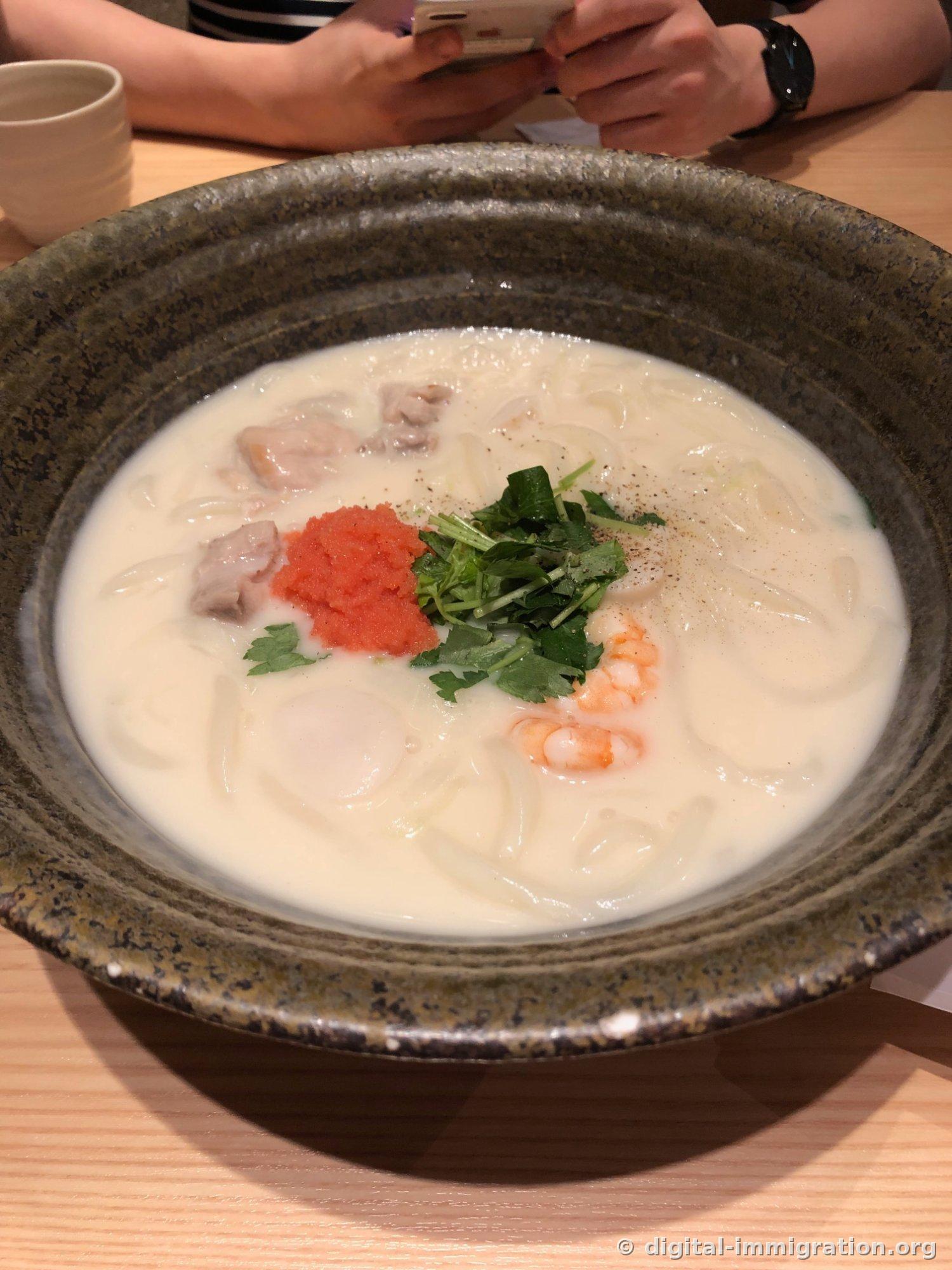 White cream udon in a large sage-colored ceramic bowl, with cod roe and mitsuba on top and chicken, scallops, and shrimp submerged in the broth