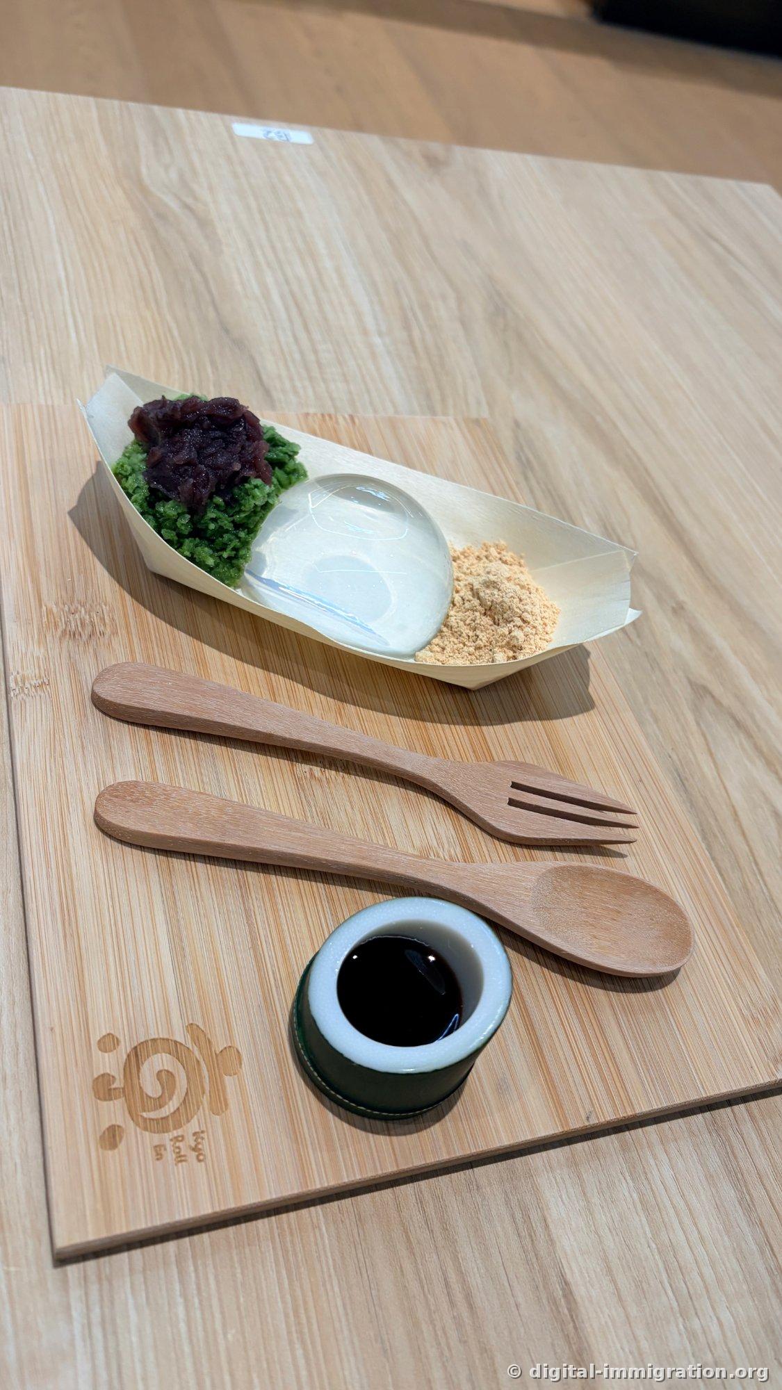 Wooden pear-shaped plate with green matcha shaved ice topped with red bean paste, transparent water drop mochi, and soybean powder arranged in a row, with bamboo fork and spoon and a small dish of black sugar syrup on a wooden placemat below