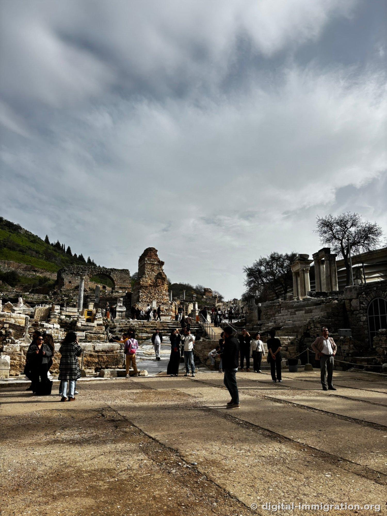 A broad paved square under cloudy skies with people scattered across it, collapsed arches and columns behind, and a modern protective roof on the right
