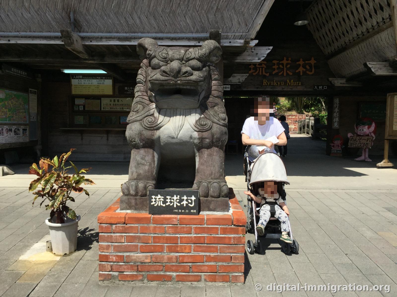 Devant l'entrée du Ryukyu Mura : un grand lion Shisa en pierre, le panneau des tarifs, et une poussette garée par un visiteur
