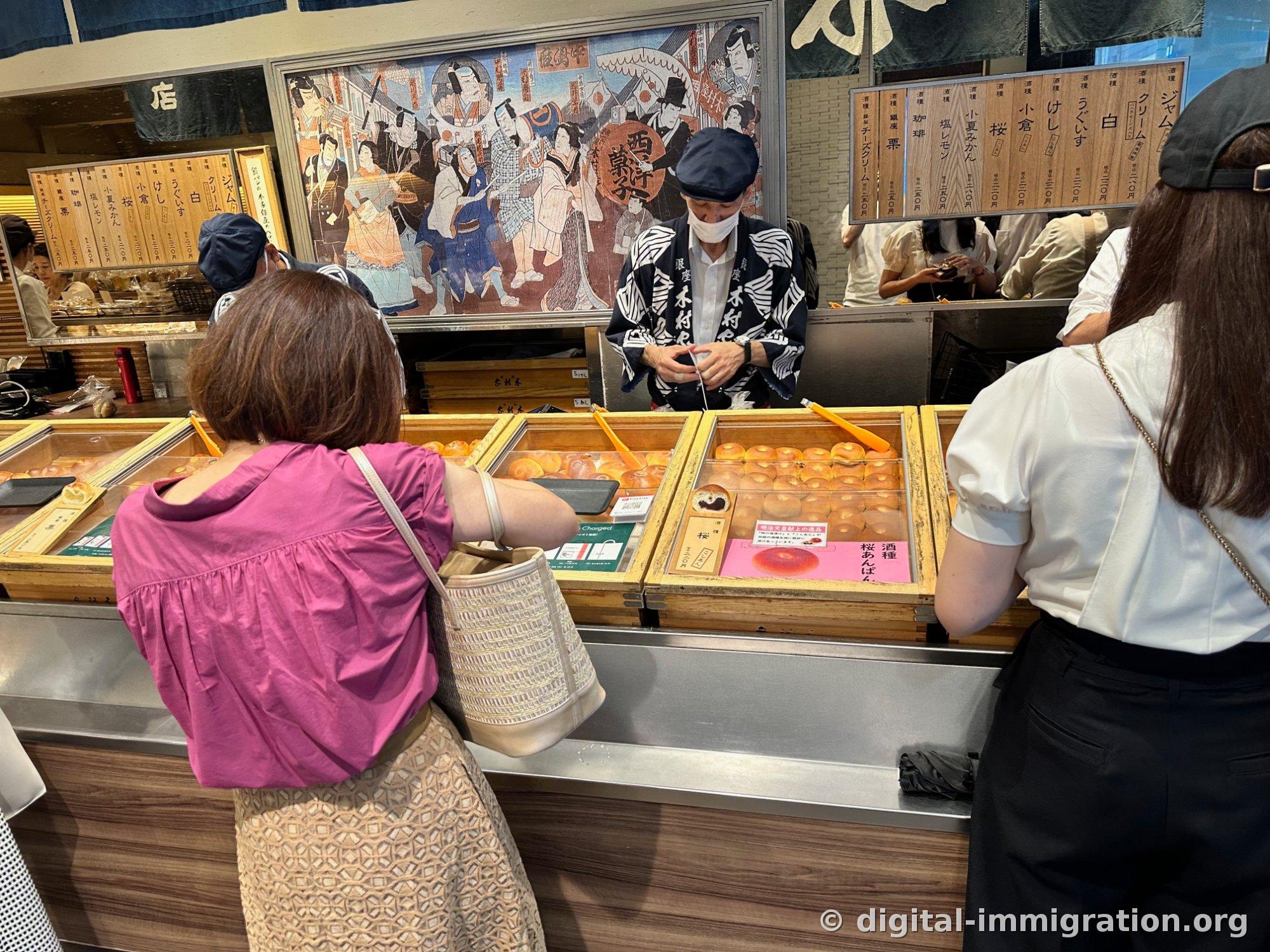 Les anpan ronds contenus dans des boîtes en bois et un employé en hapi bleu-gris se tenant derrière, avec des étiquettes de prix en bois par type et des gravures ukiyo-e accrochées au mur