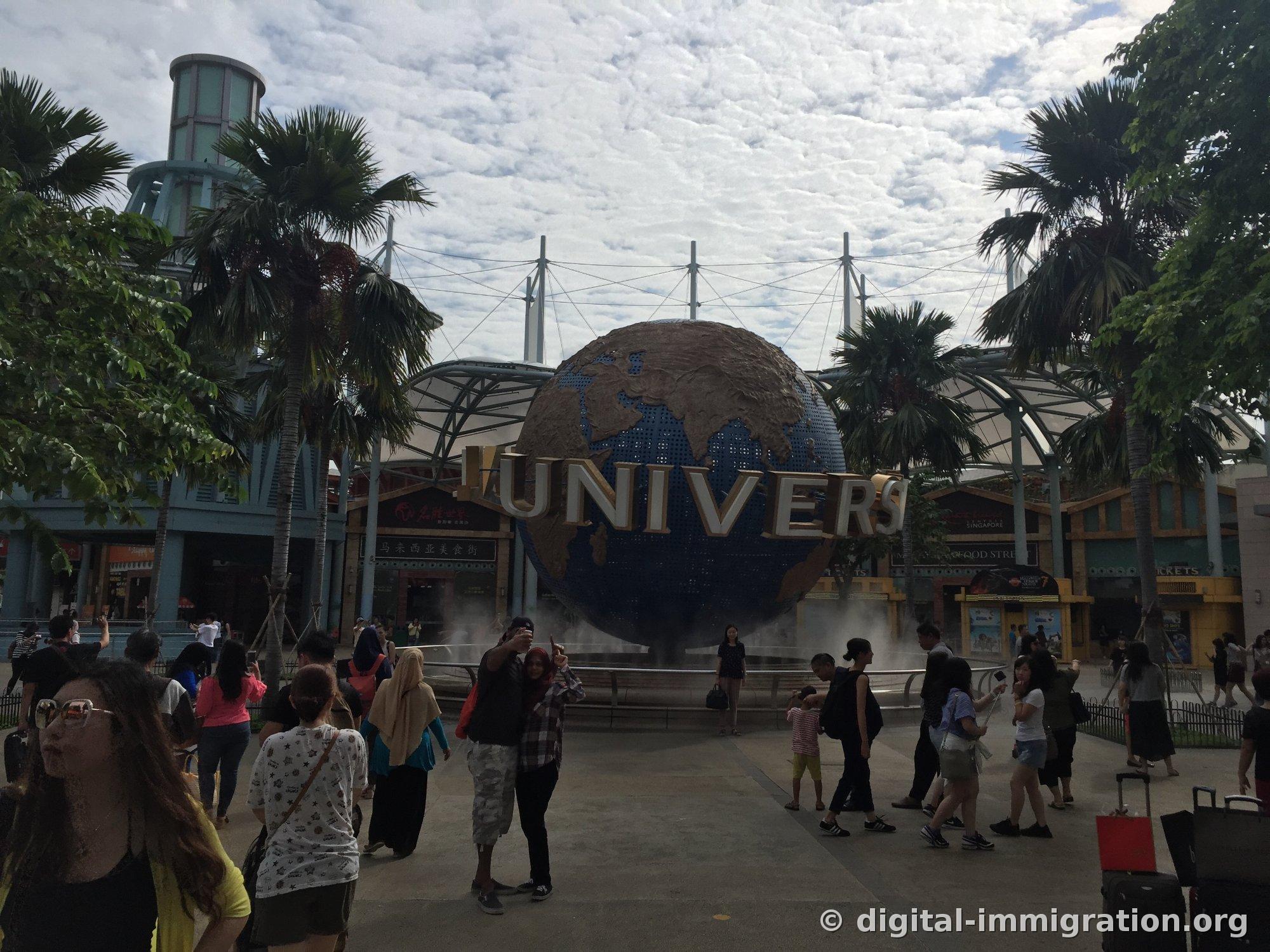 Tourists taking photos in front of the rotating globe at the main entrance of Universal Studios Singapore under palm trees and cloudy sky