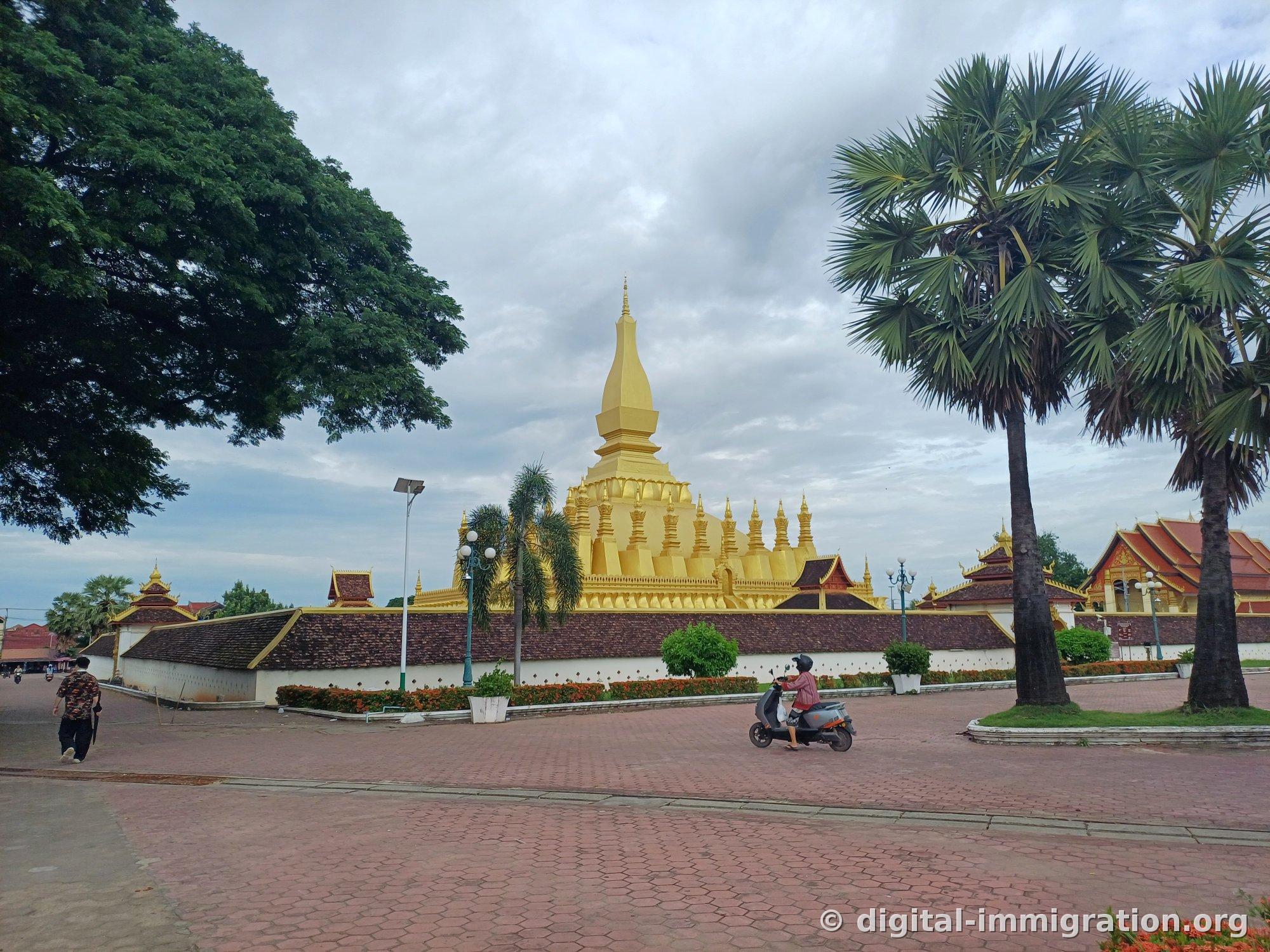 Pha That Luang viewed from the red brick plaza with palm trees and a person on a motorcycle passing by