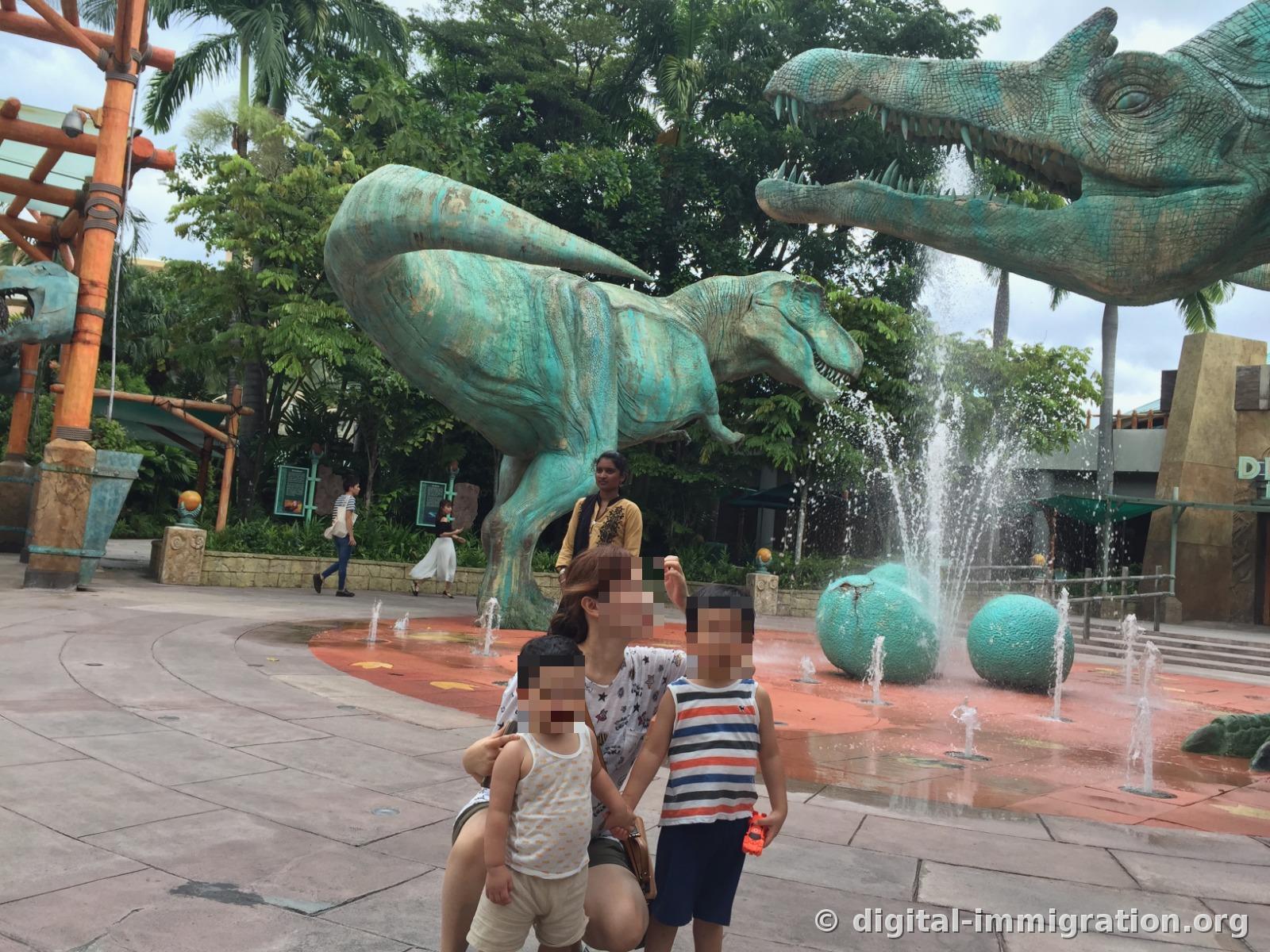 A family sitting in front of the turquoise Tyrannosaurus dinosaur sculpture and fountain in the Jurassic Park water play plaza