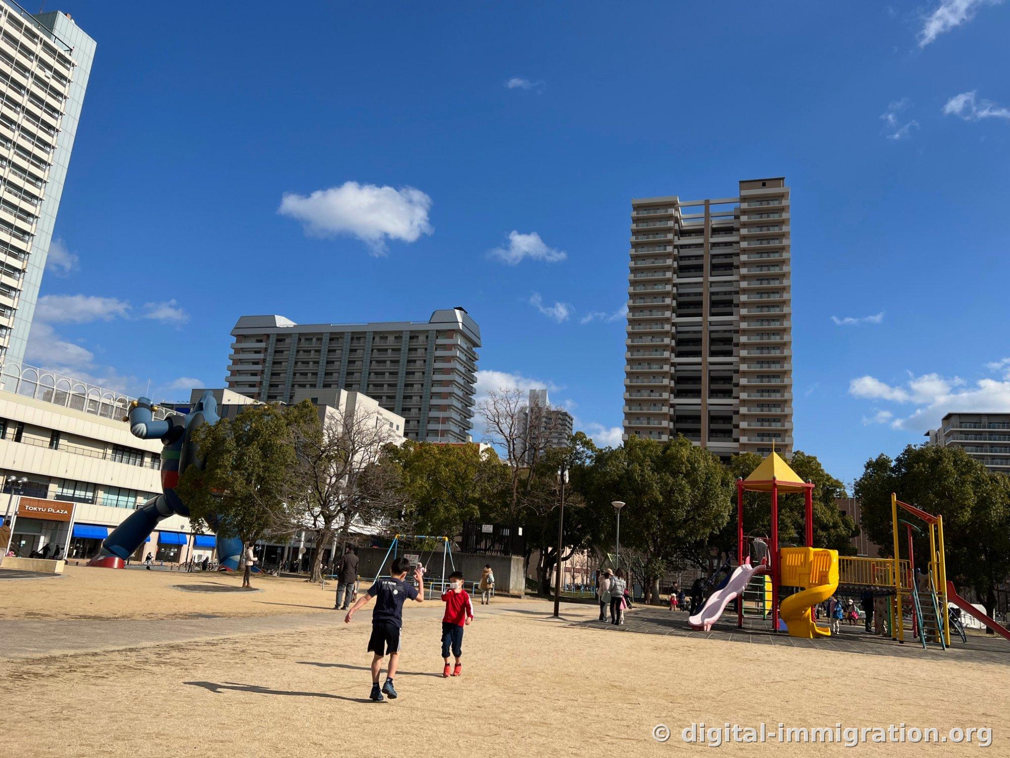 Deux enfants courant dans une vaste esplanade en terre battue, avec un toboggan jaune sur la droite et les jambes de Tetsujin 28 visibles en bas à gauche