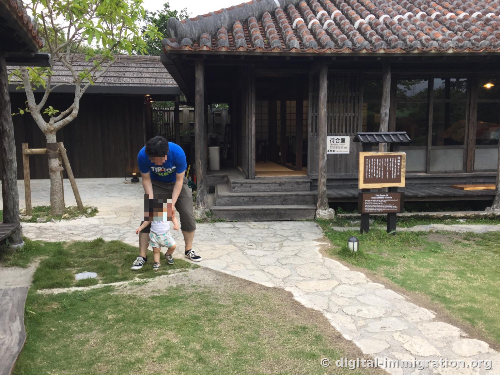붉은 류큐 기와지붕의 목조 건물 앞 잔디밭에서 어른이 아기 손을 잡고 걸음마를 시키는 모습, 오른쪽에 '待合室 Waiting room' 안내판
