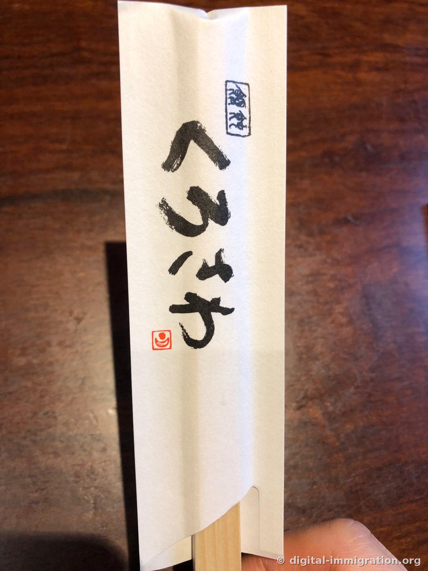 筆書きで くろさわ と書かれ、赤い落款が押された白い箸袋を手に持っている様子