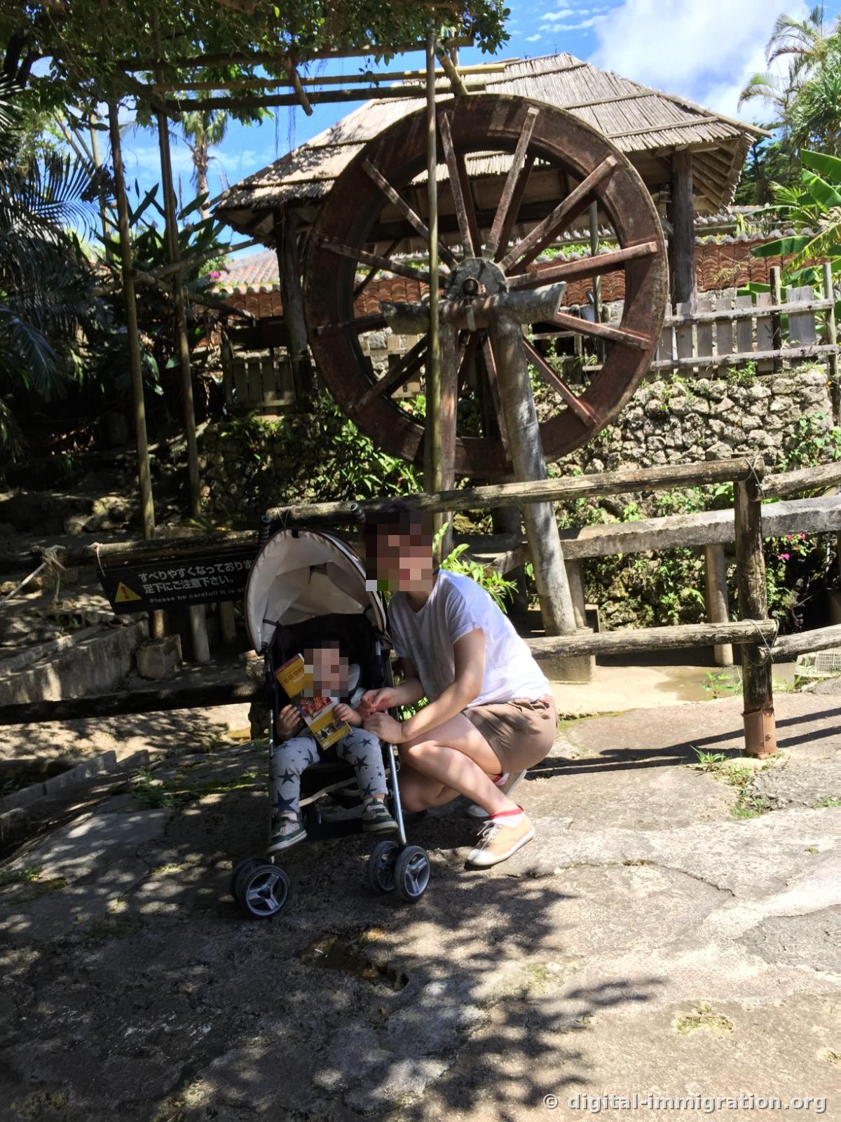 Devant un grand moulin à canne à sucre avec une roue de bois couverte d'un toit, un visiteur est assis avec une poussette, avec un panneau d'avertissement « Attention au sol glissant » à côté