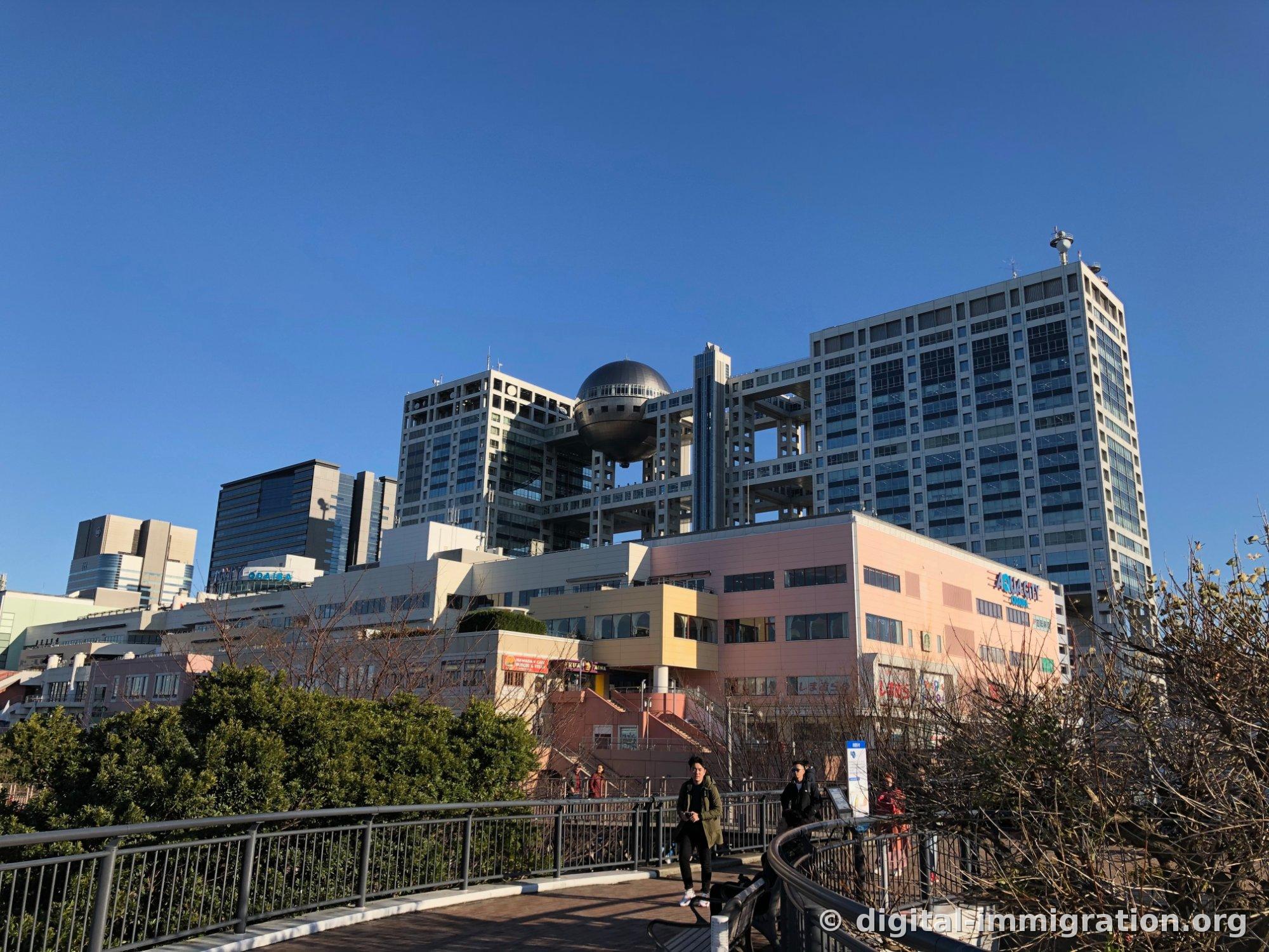 从台场天桥仰视富士电视本社建筑的银色球形瞭望台，前面是粉红色AquaCity台场商业建筑