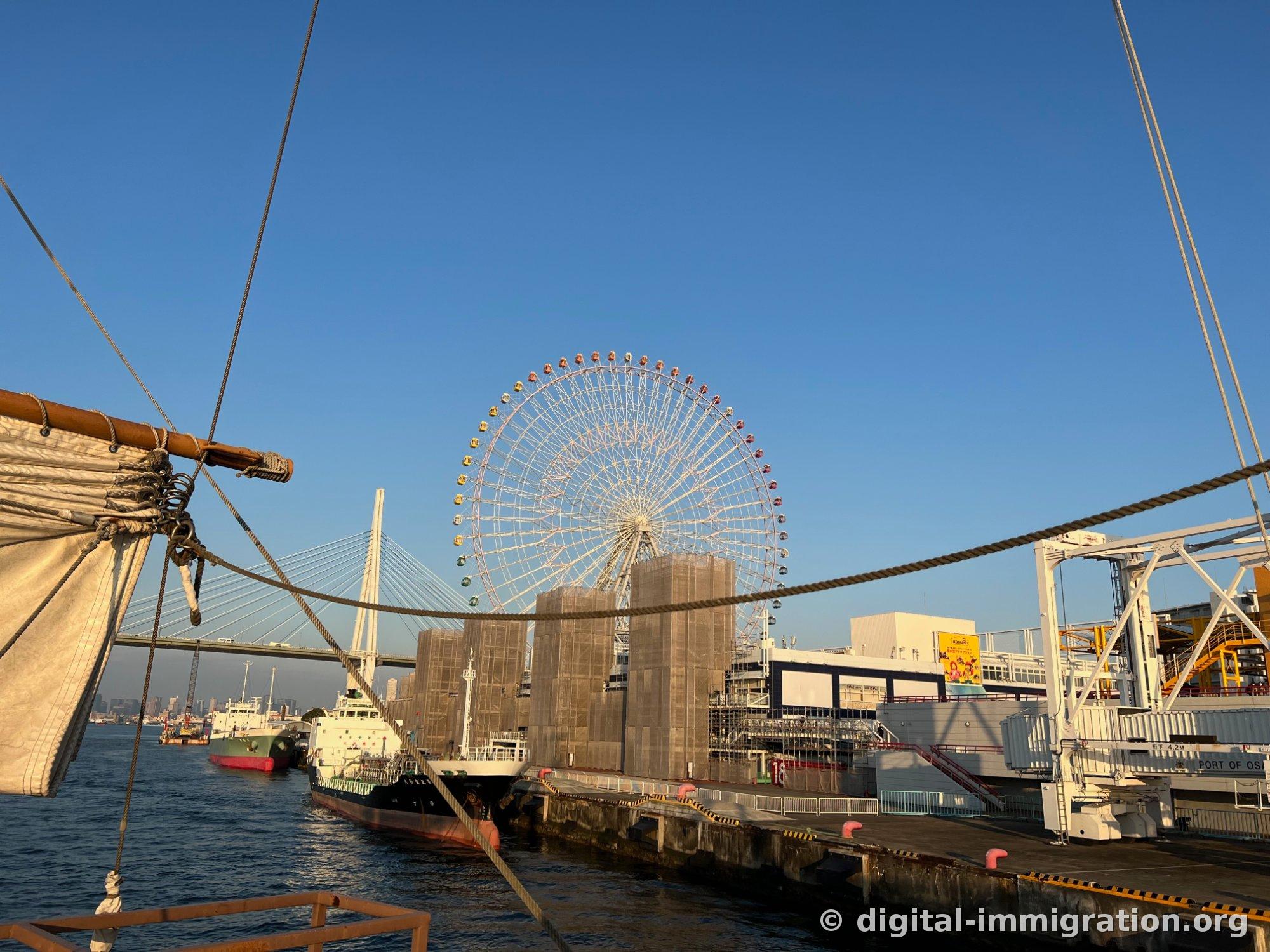 サンタマリア号の帆と太いロープの向こうに、カラフルなゴンドラの天保山大観覧車と白い斜張橋が見える港の風景