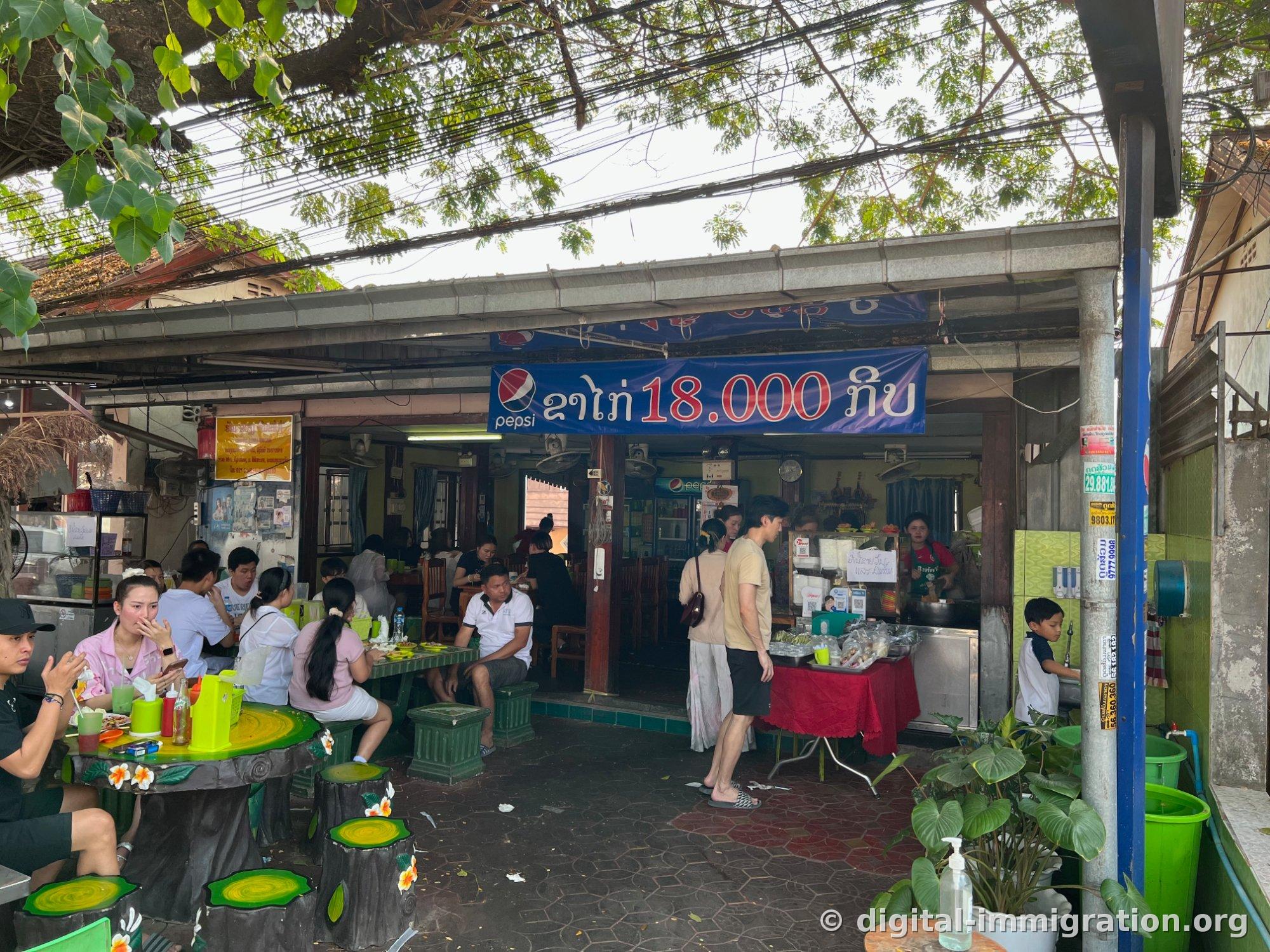 The shop entrance with the blue Pepsi banner reading 'chicken 18,000 kip' and the order counter covered with red cloth, with customers eating while sitting on wooden stump-shaped chairs
