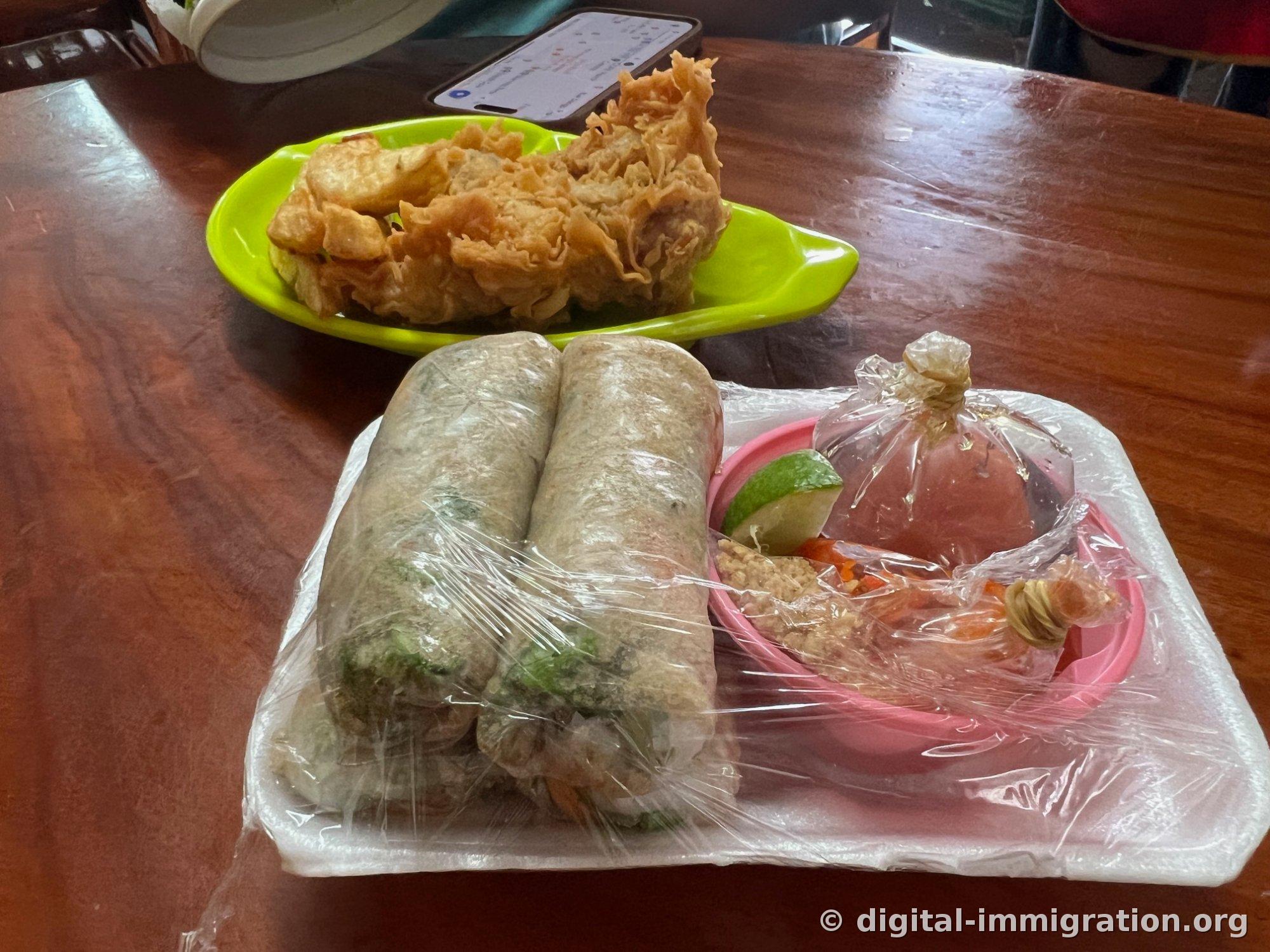 Fried items and potatoes on a pale-green plate on a wooden table, with two rice paper rolls wrapped in plastic, lime, peanuts, and sauce in bags on a pink plate in the foreground