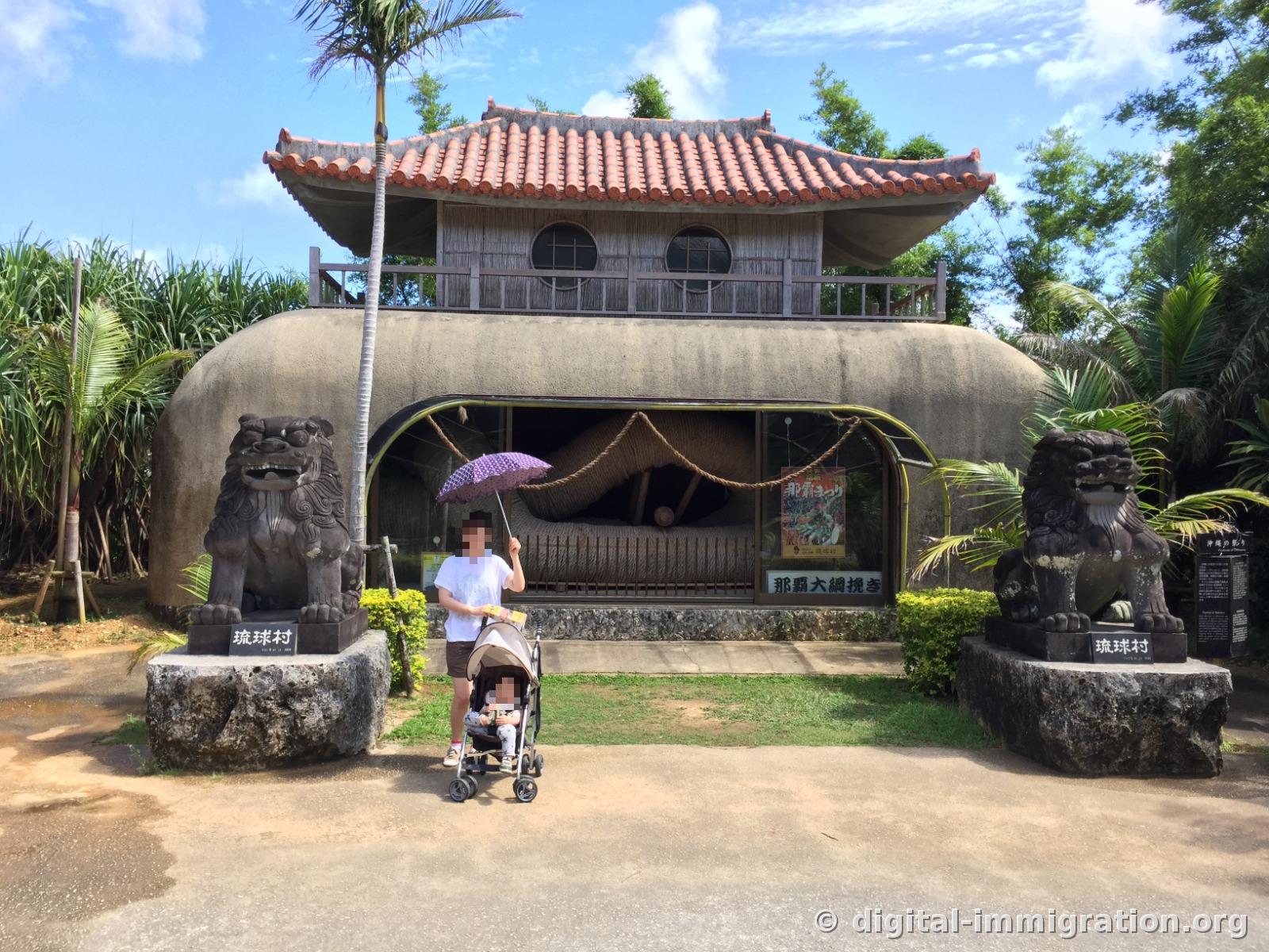 Une personne avec un parasol violet est debout avec une poussette devant l'exposition du grand tir à la corde de Naha au Ryukyu Mura