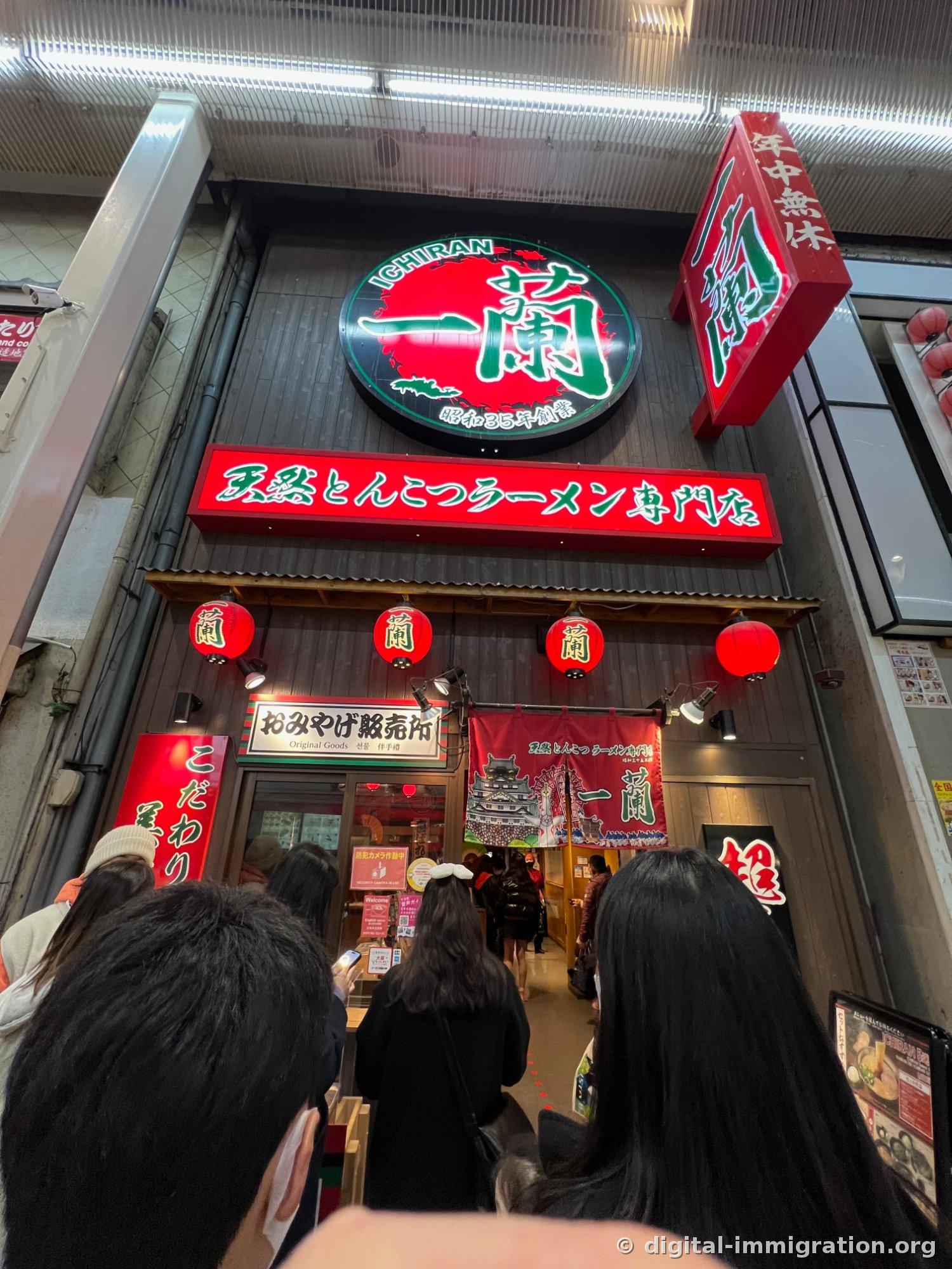 The entrance to Ichiran Dotonbori Annex visible atop the arcade stairs, with its green-bordered circular sign, red lanterns, and people waiting in line