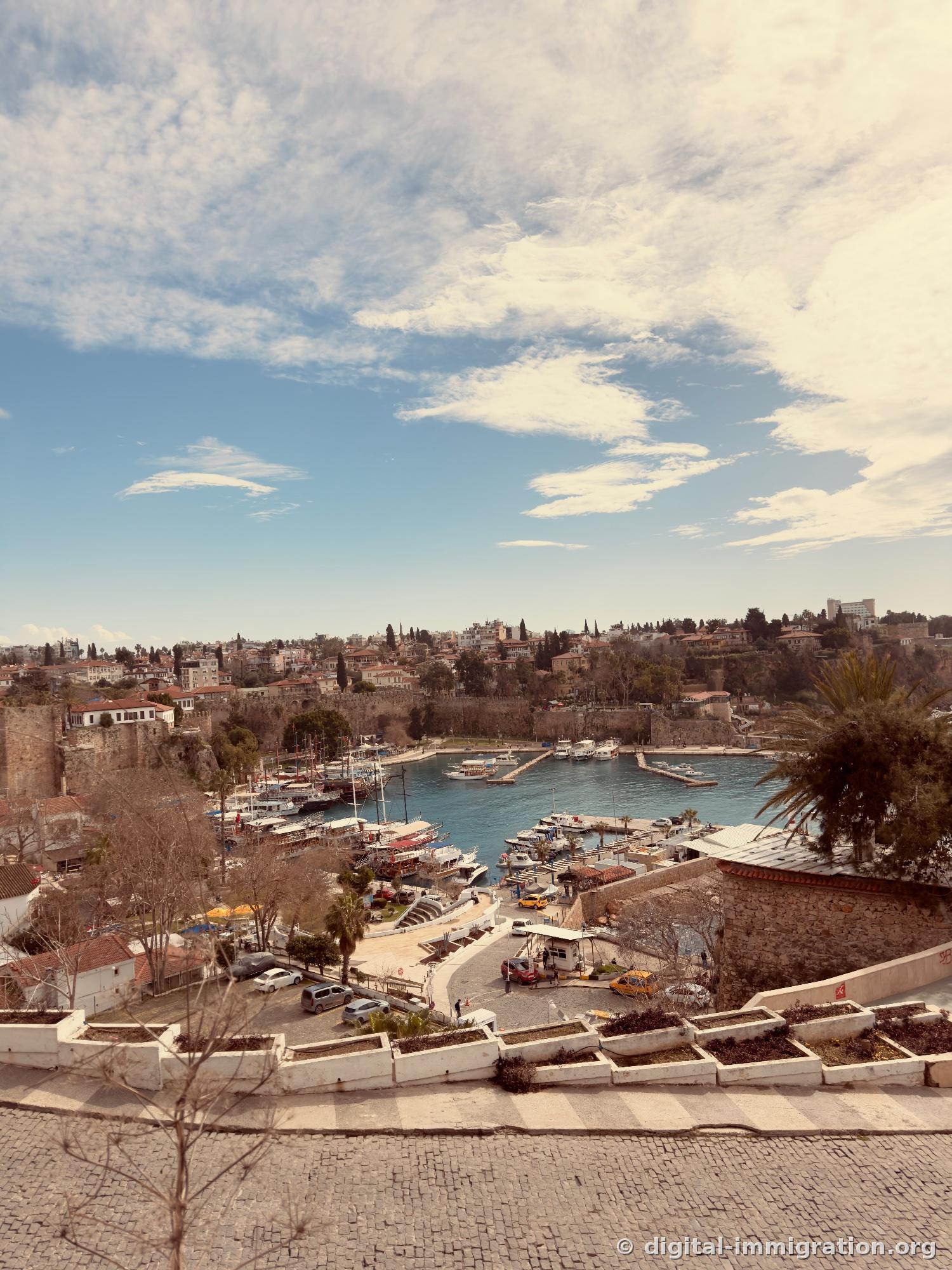 A sweeping view of Antalya's yacht harbor from the cliff above Kaleiçi, with city walls, red-tiled roofed houses, moored wooden boats, and turquoise sea visible below