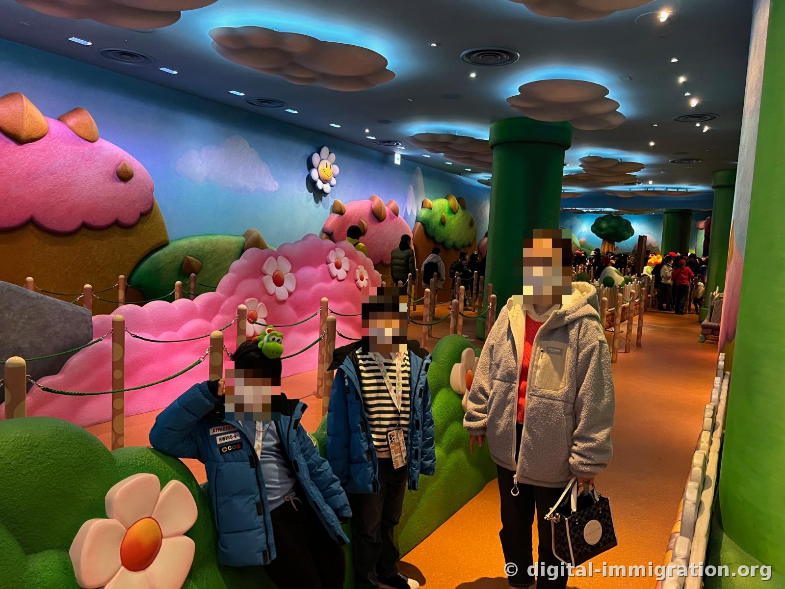 Yoshi Adventure indoor queue corridor. A family of three stands under pink hills, white flower sculptures, and three-dimensional clouds on the ceiling