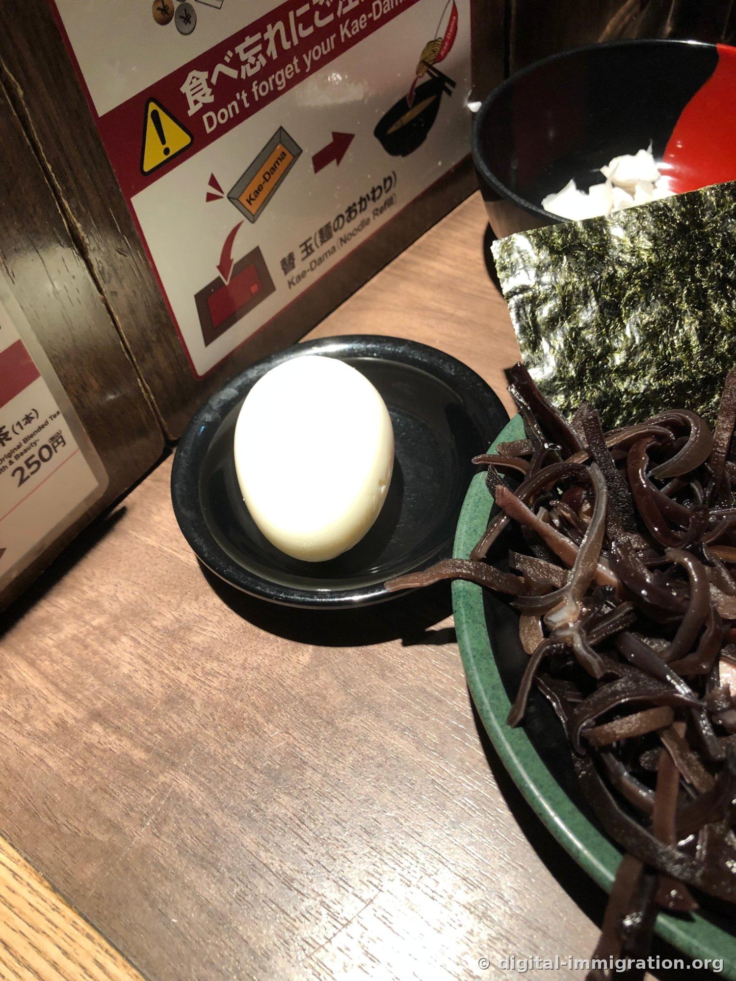 Un œuf bouilli sans coquille dans un petit bol noir, avec un panneau de rappel « N'oubliez pas le kaedama » derrière