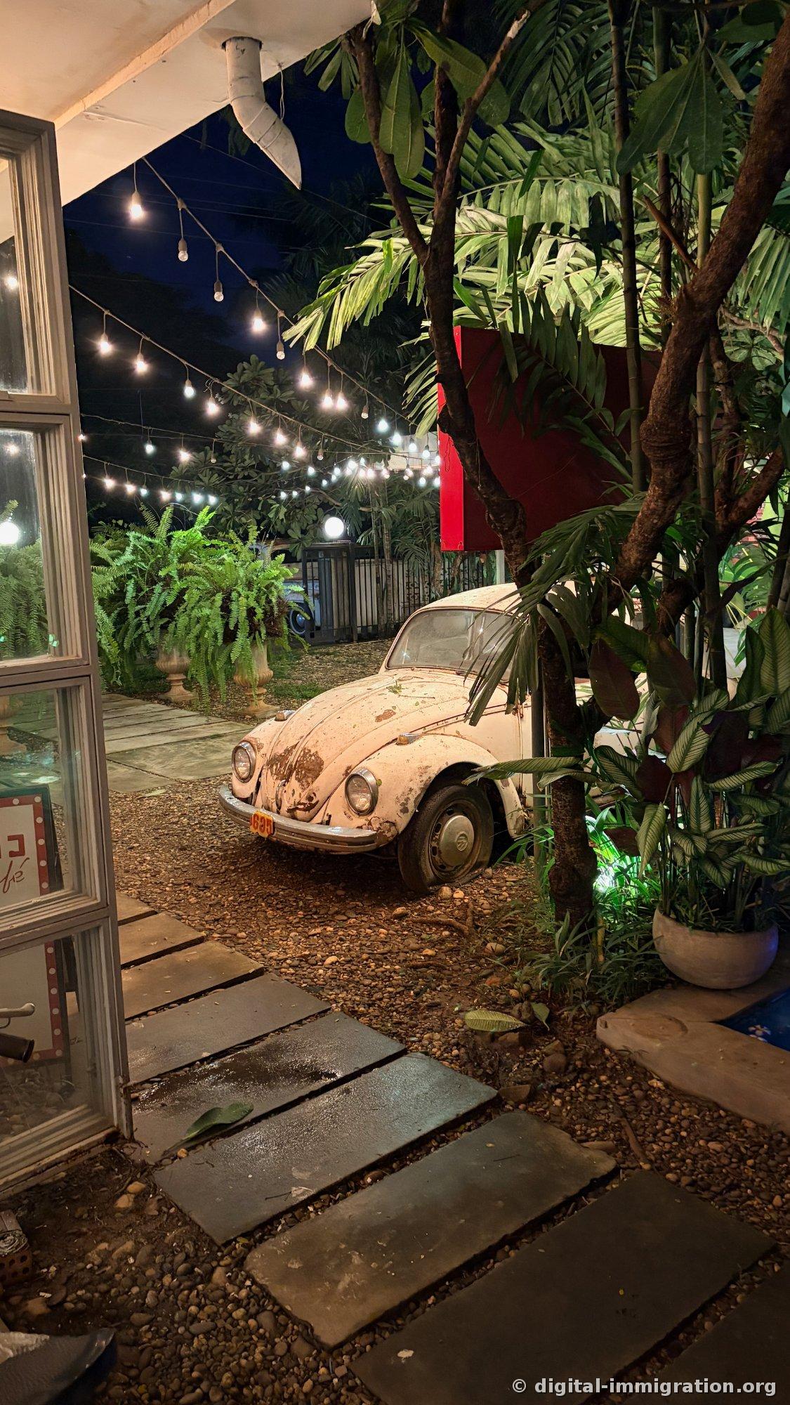 Une vue du jardin la nuit à travers une porte vitrée depuis l'intérieur, la Coccinelle blanche sous les guirlandes lumineuses et des marches en pierre sur le gravier