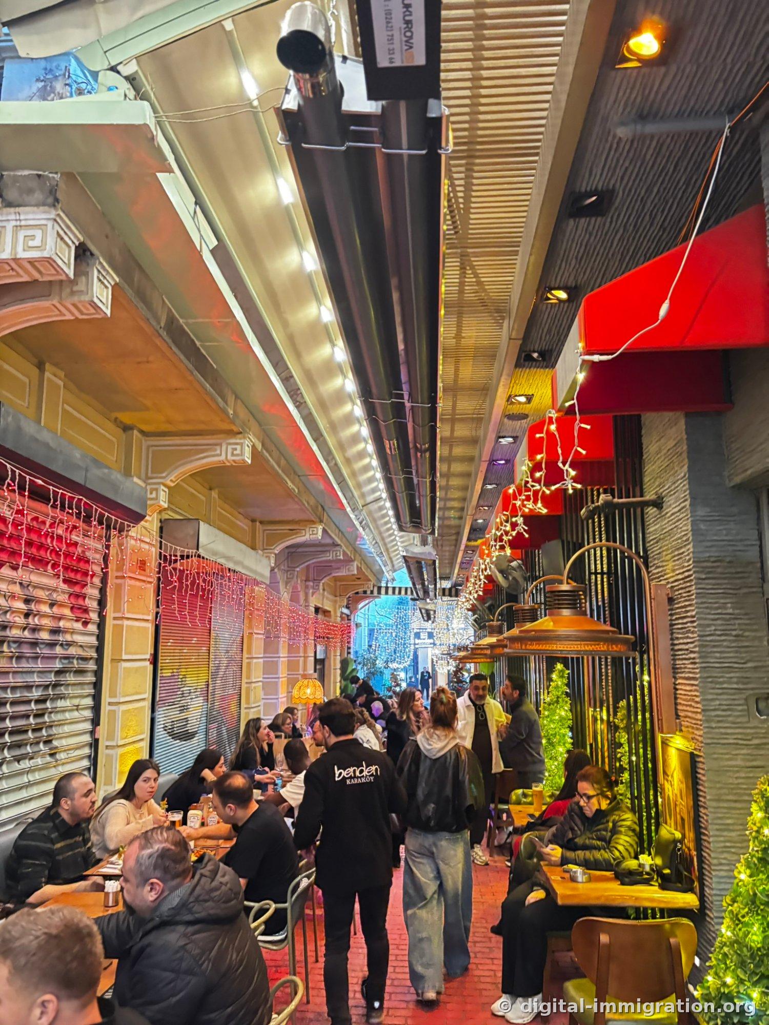 Narrow alley in Karaköy with heaters and light decorations hanging, people sitting at tables on both sides, and a staff member in a benden KARAKÖY uniform passing by