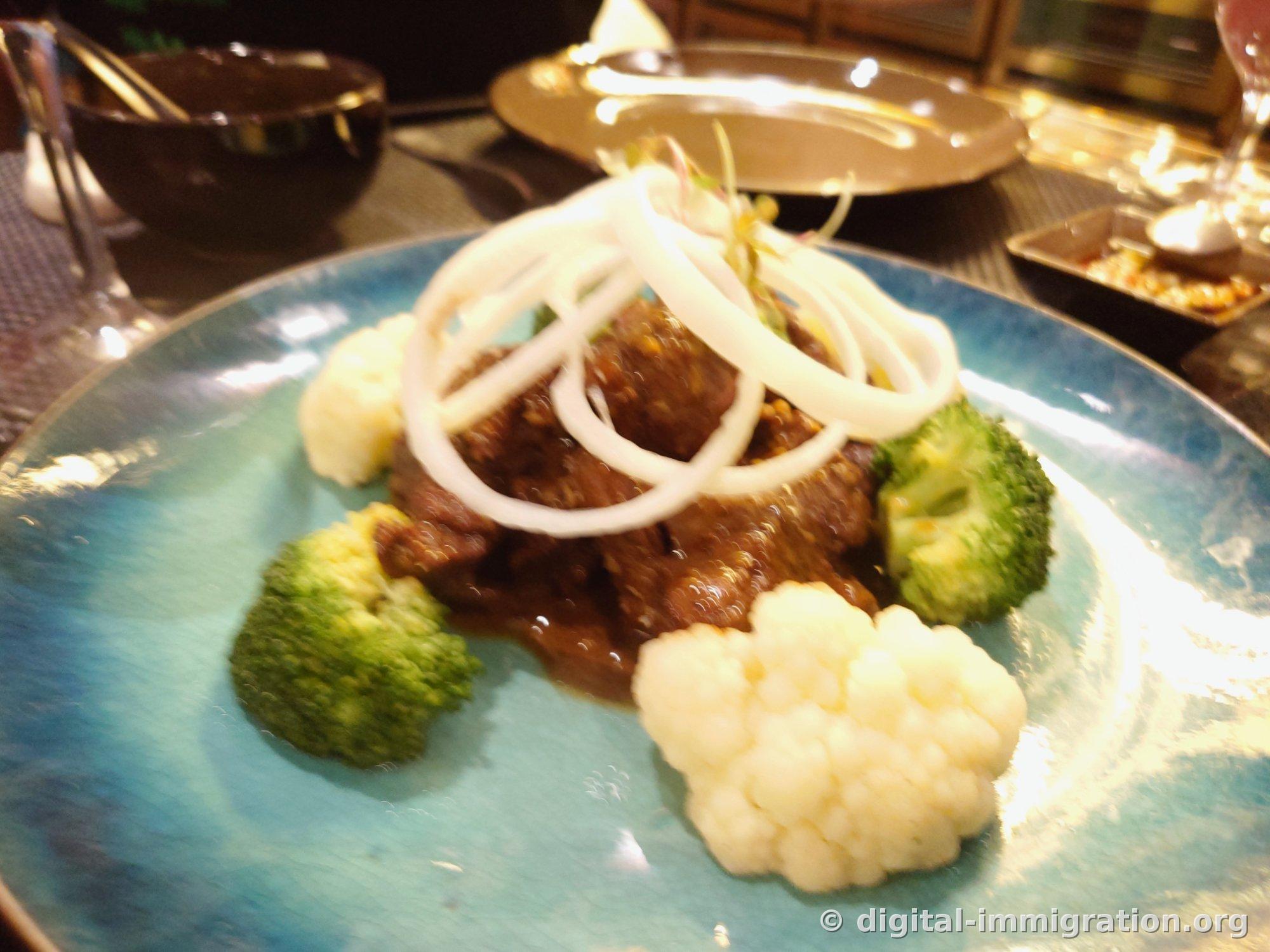 Beef stew in brown sauce on a blue plate topped with raw onion rings, served with broccoli and cauliflower