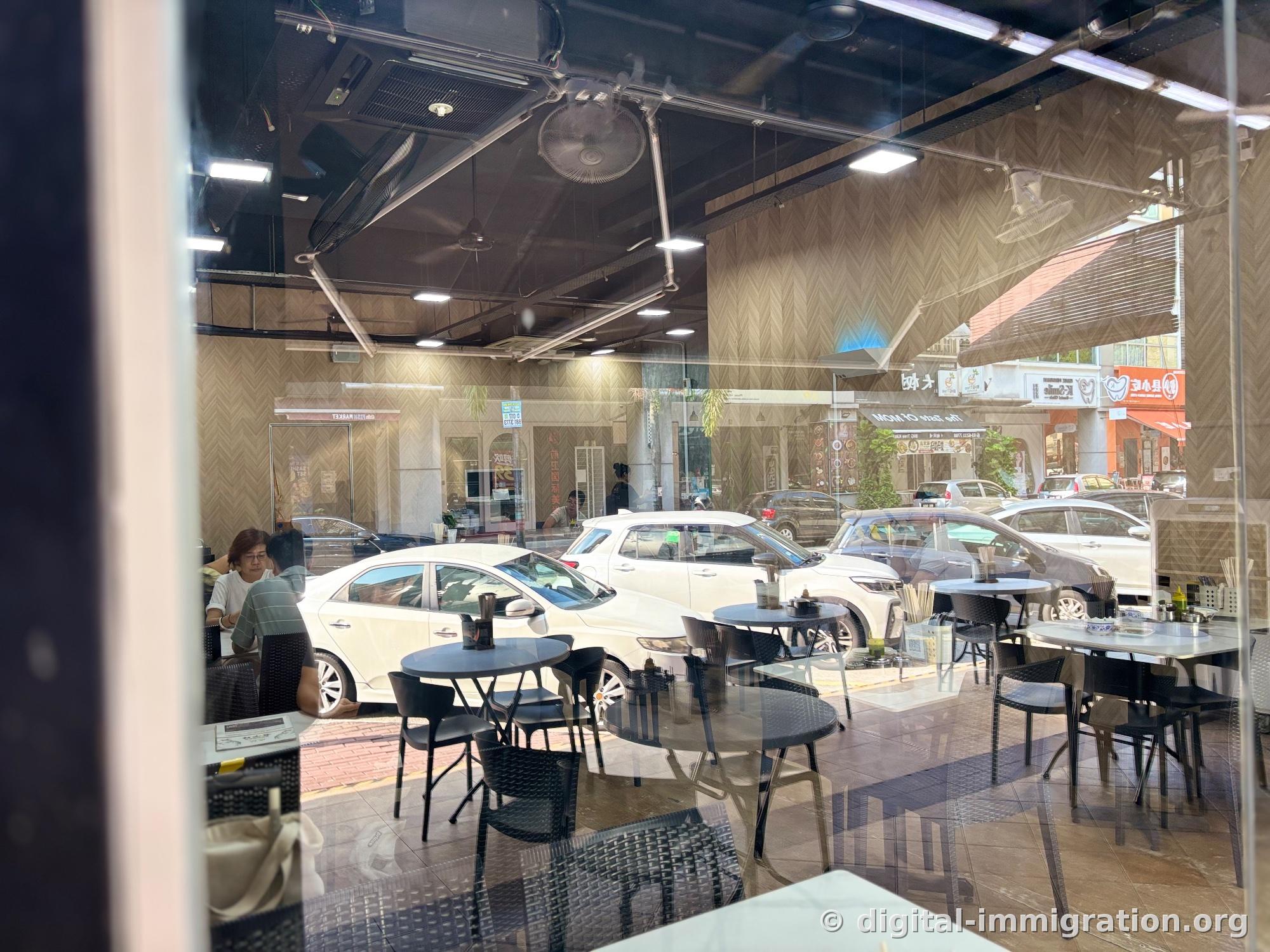 View through the restaurant's interior glass window showing the outside. White parked cars and shop signs across the street reflect and overlap on the glass