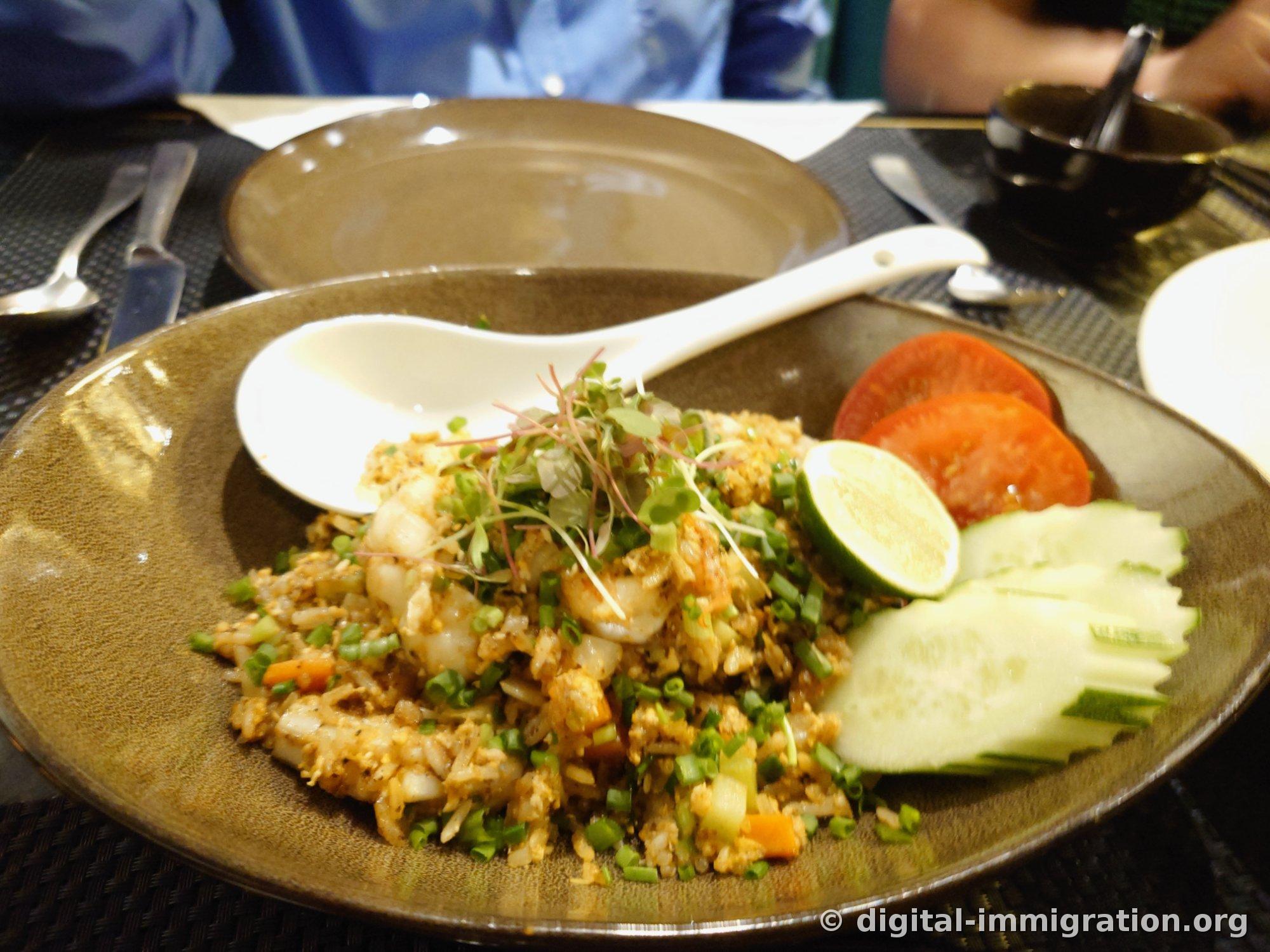 Shrimp fried rice on a brown ceramic plate topped with scallions and microgreens, served with lime, tomato, and cucumber