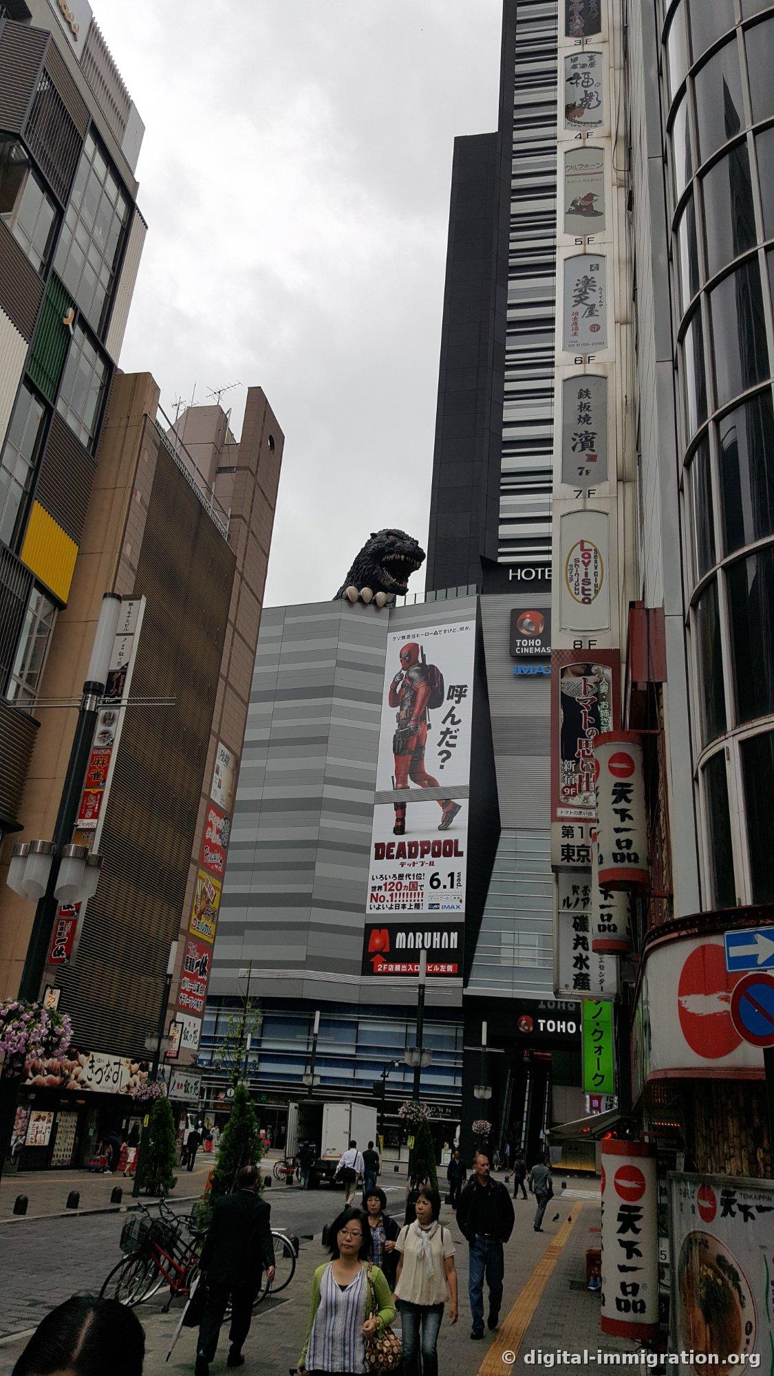 La rue de Kabukicho par temps couvert. La tête de Godzilla sur le bâtiment avec la grande affiche Deadpool, l'enseigne TOHO CINEMAS, les enseignes des magasins en japonais alignées verticalement, et les piétons qui passent
