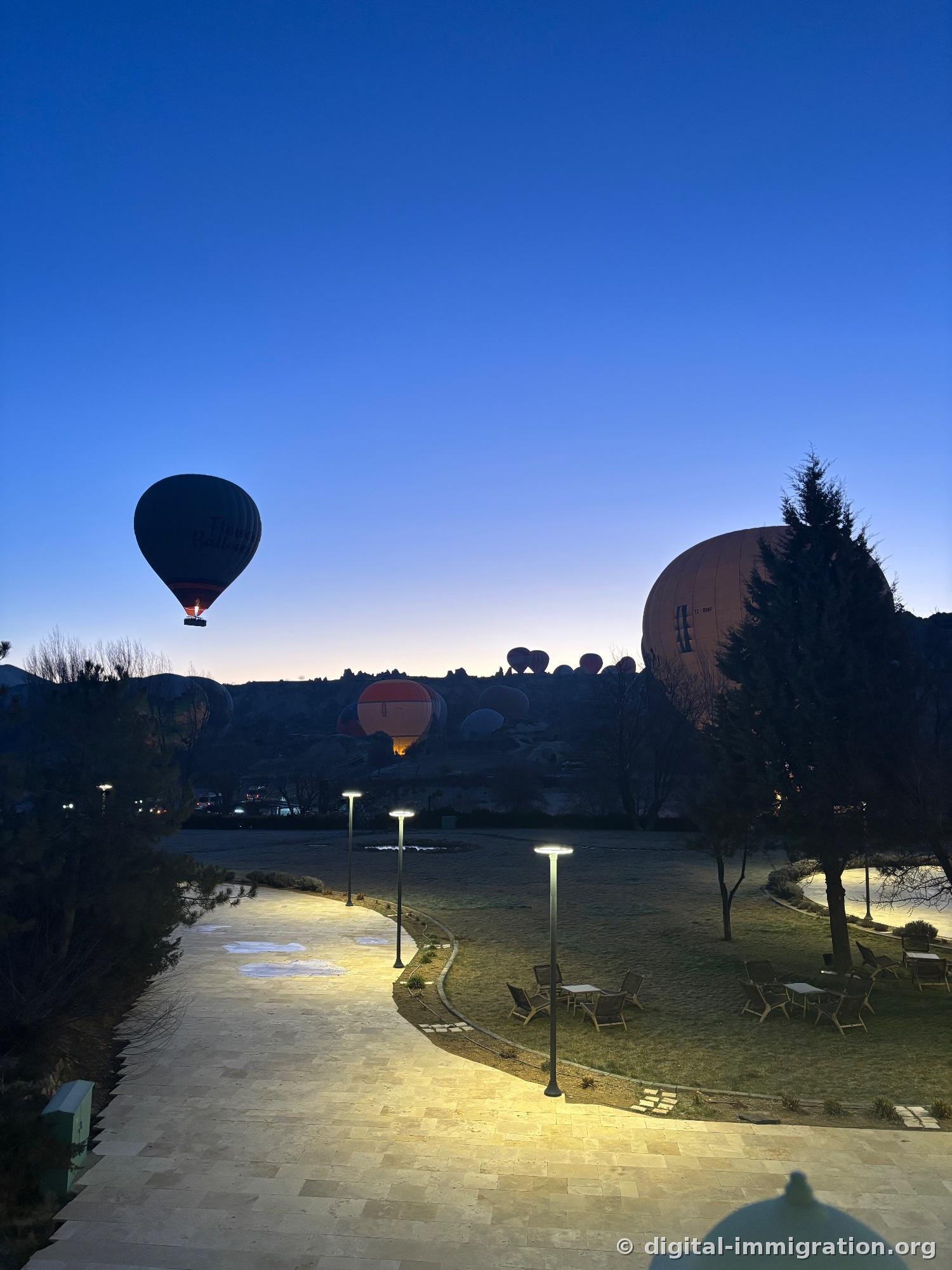 Une montgolfière volant dans un ciel bleu pâle à l'aube, et à droite derrière les arbres, une énorme montgolfière gonflée, en bas le sentier du jardin de l'hôtel éclairé et les tables extérieures