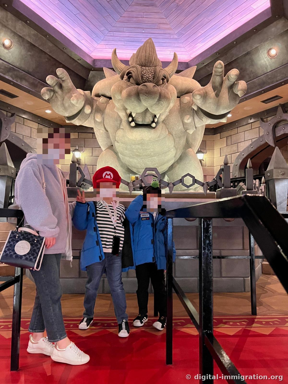 Children and an adult wearing Mario and Yoshi hats pose for a photo in front of a giant gray Bowser statue inside Bowser's Castle