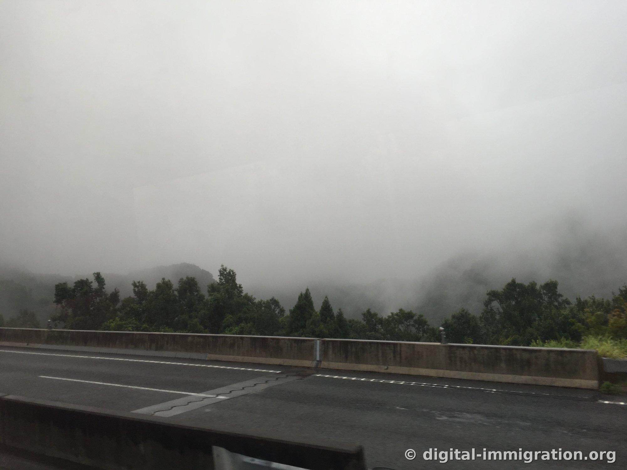 Un mur antibruit d'autoroute visible par la fenêtre de la voiture et une montagne enveloppée de nuages, avec l'asphalte mouillé