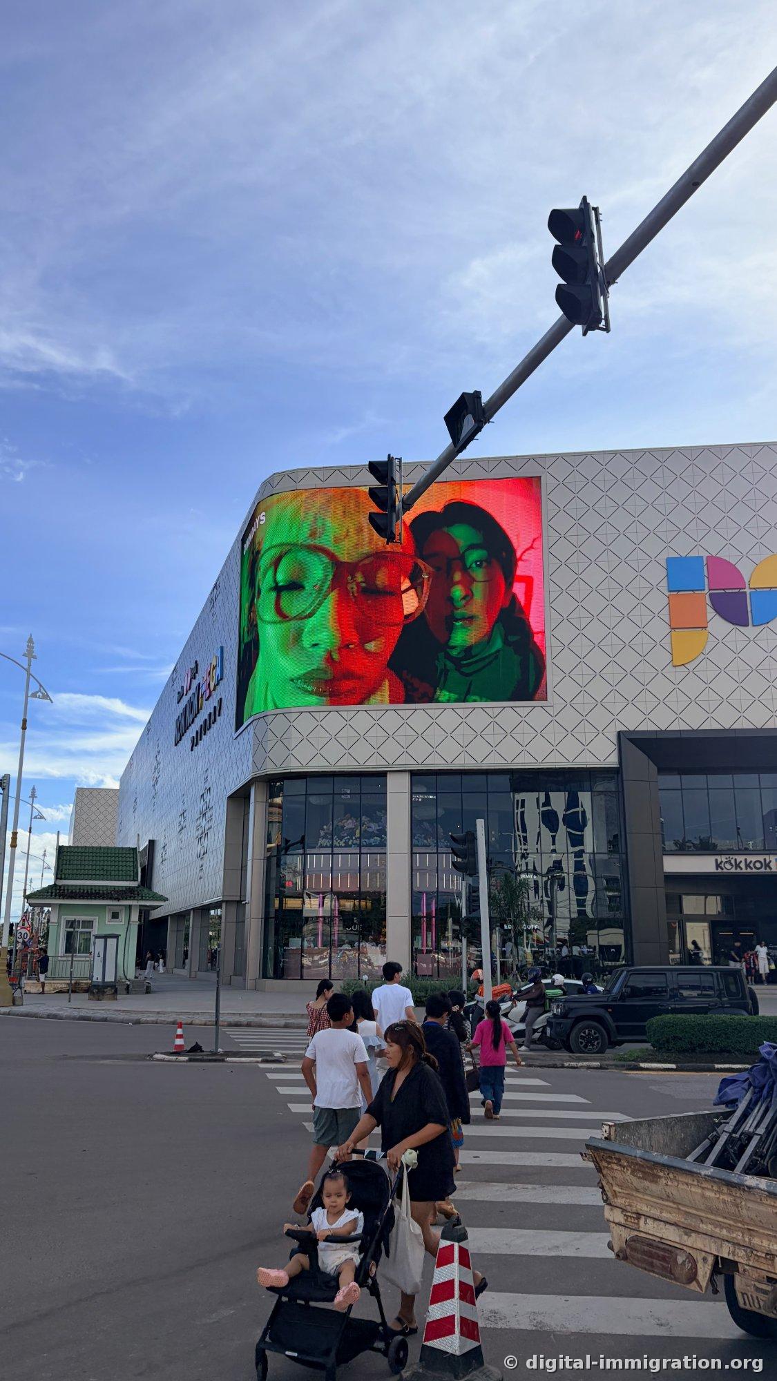 Kokkok Mega Mall exterior viewed from across the street, curved LED screen and white triangular pattern façade, people with motorcycles and strollers