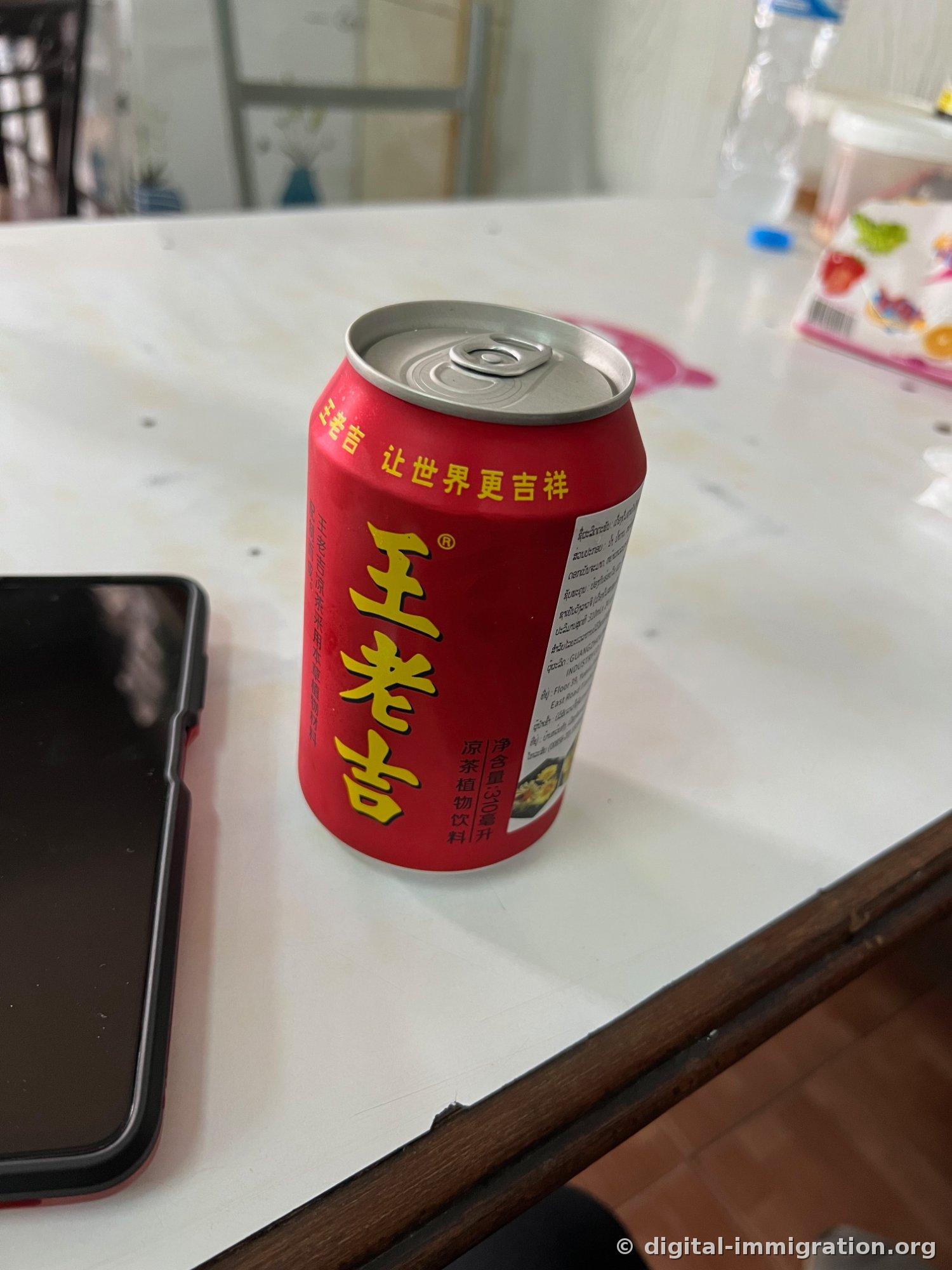 A red Wang Lao Ji herbal tea can on a white table with a Lao language ingredient label, with a smartphone beside it
