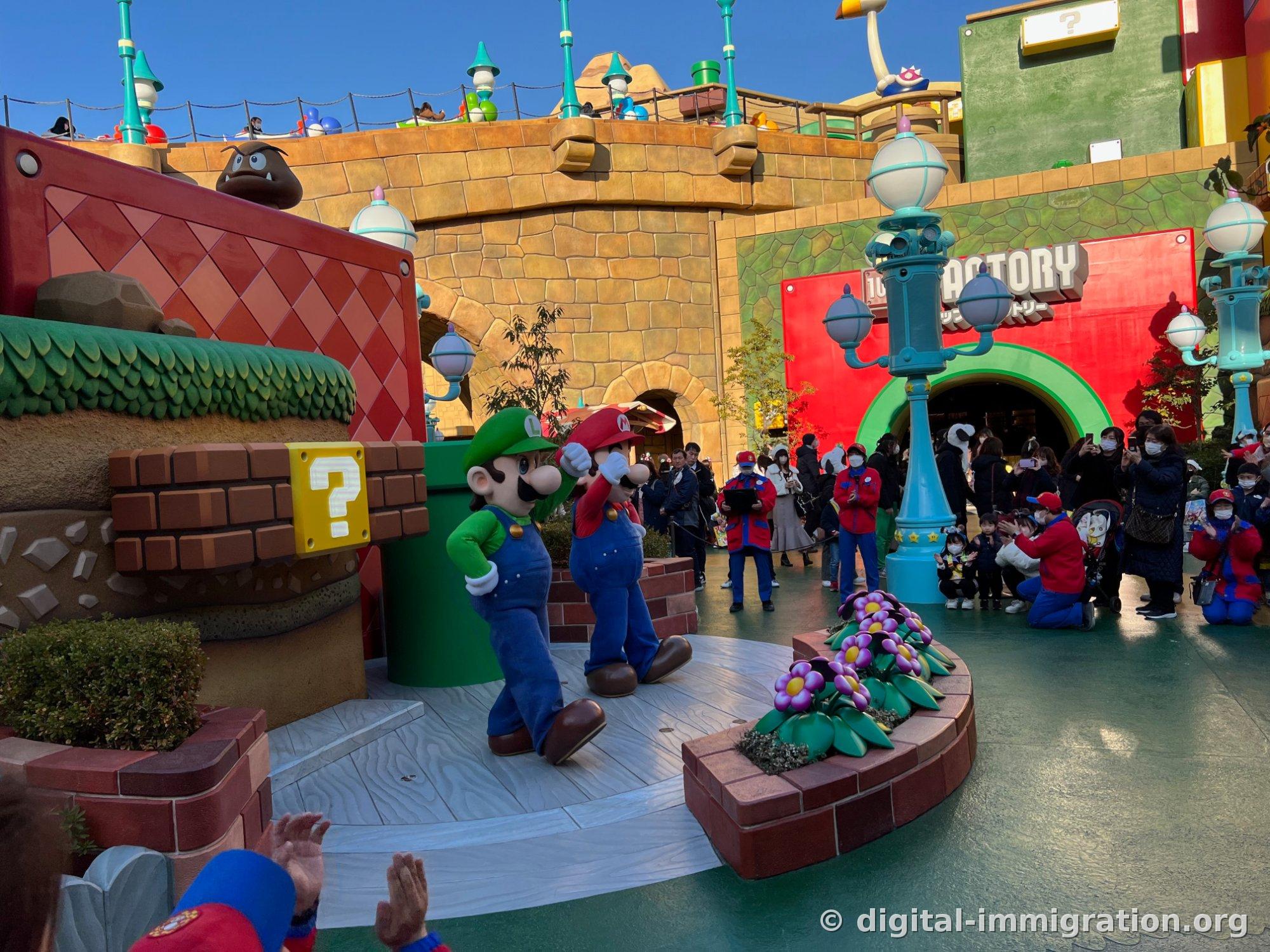 Luigi and Mario stand side by side posing on the circular stage, with the audience in front applauding