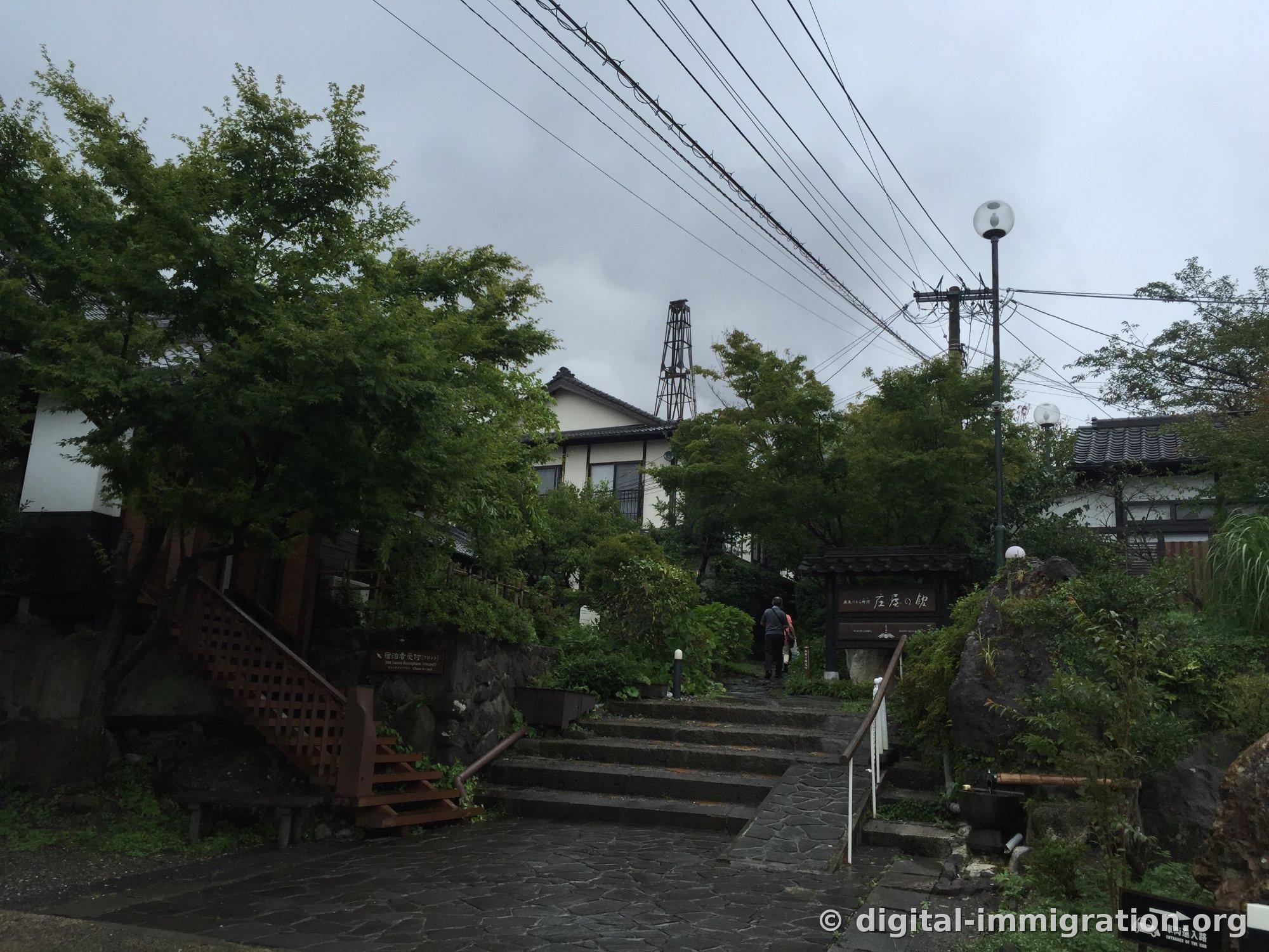 Une personne montant un escalier en pierre sous la pluie et le panneau de 庄屋の館, avec des fils téléphoniques sous un ciel couvert