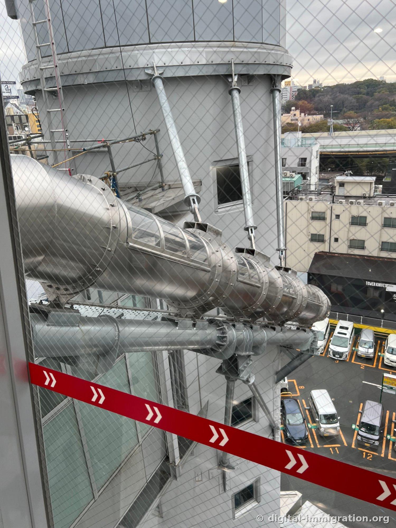 The silver spiral tube structure of the Tower Slider visible through the observation deck window, with parking lot and TOWER KNIVES OSAKA sign below