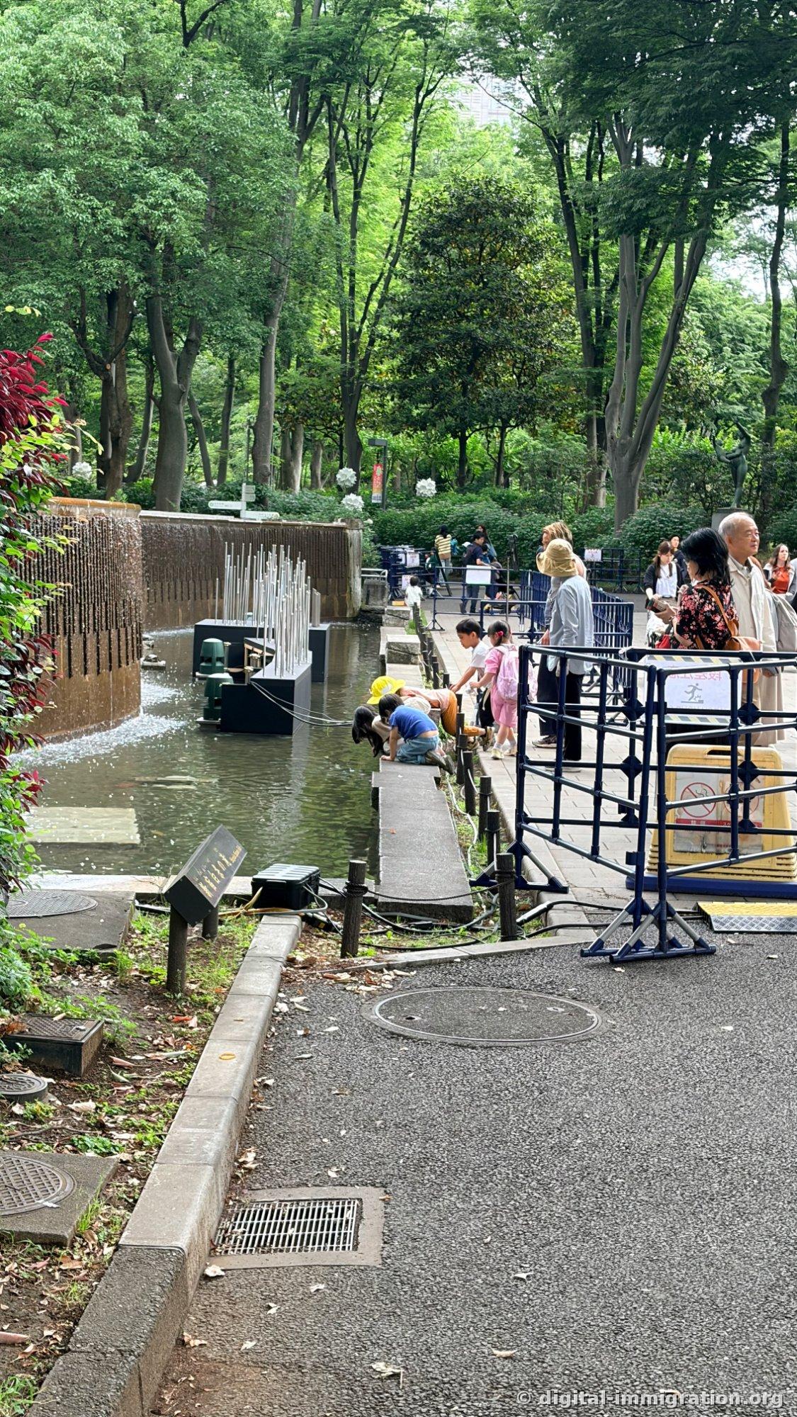 Cascade artificielle avec des enfants accroupis au bord de l'eau peu profonde regardant dedans, avec une clôture de sécurité bleue à l'arrière et des adultes surveillants