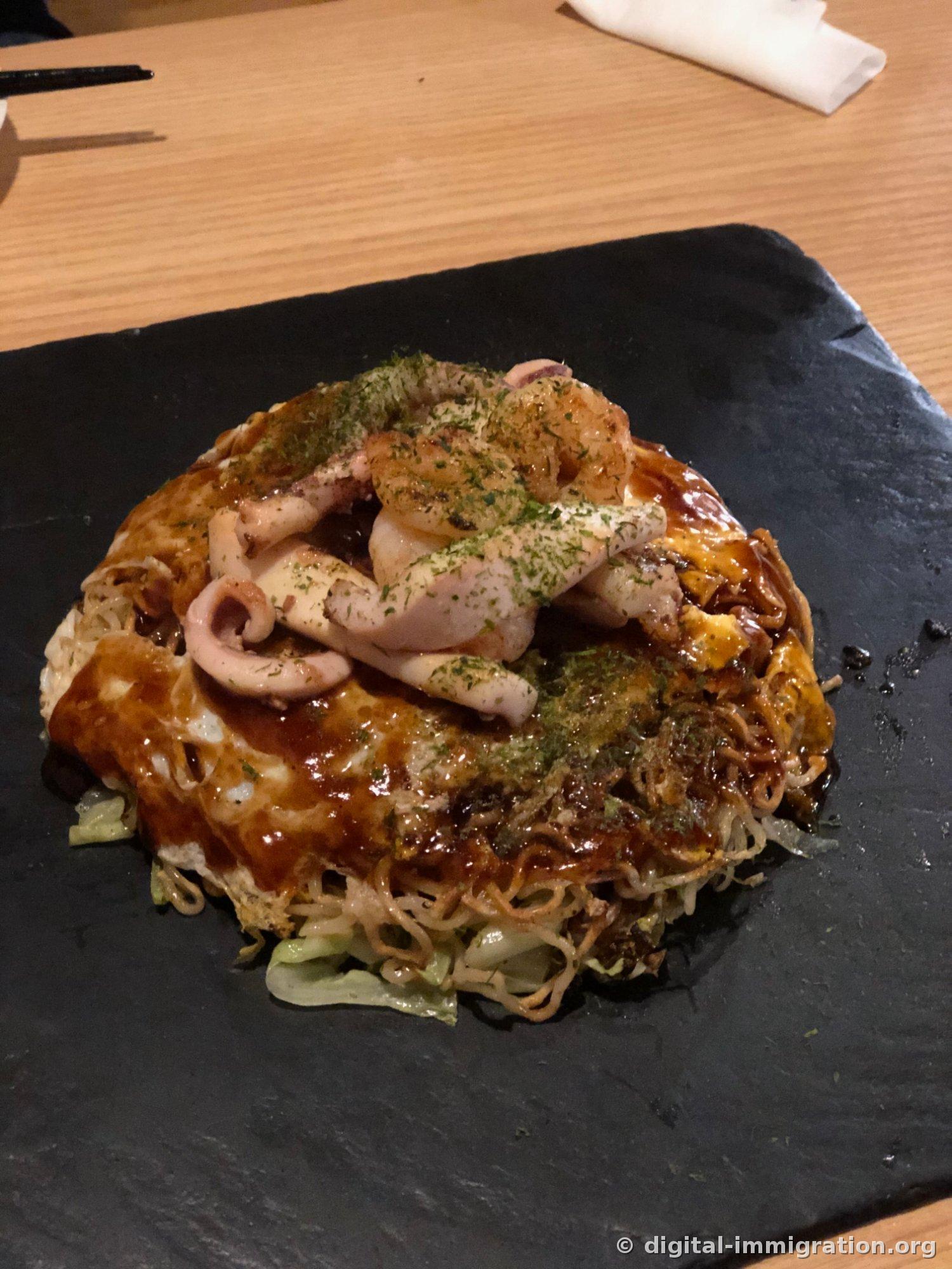 검은 슬레이트 접시에 담긴 히로시마식 오꼬노미야끼, 위에 오징어와 아오노리, 옆으로 노릇하게 구워진 야끼소바 면이 보인다