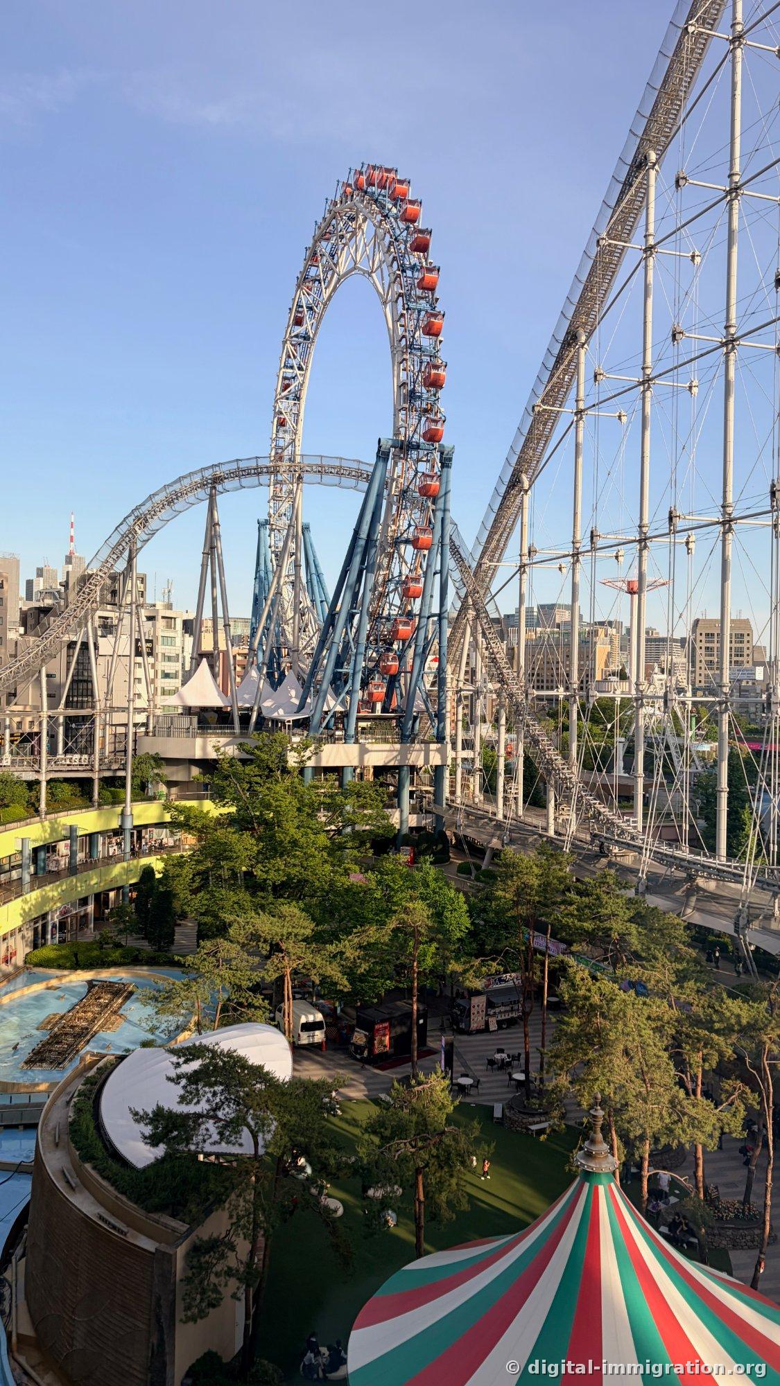 縦構図で見下ろした東京ドームシティ全景、観覧車とローラーコースター下の緑の芝生と赤緑の縞模様テント、青いウォーターアトラクション