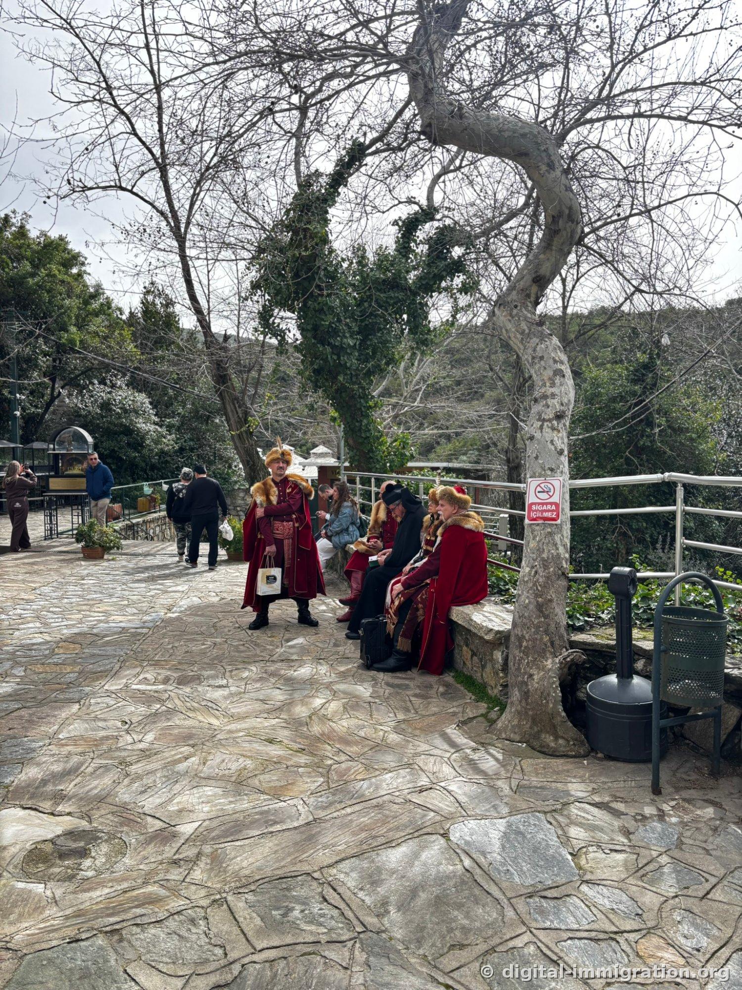 People in crimson velvet traditional dress trimmed with fur and feathered hats stand or sit on benches in a stone plaza, with a red SIGARA ICILMEZ (NO SMOKING) sign on the nearby railing
