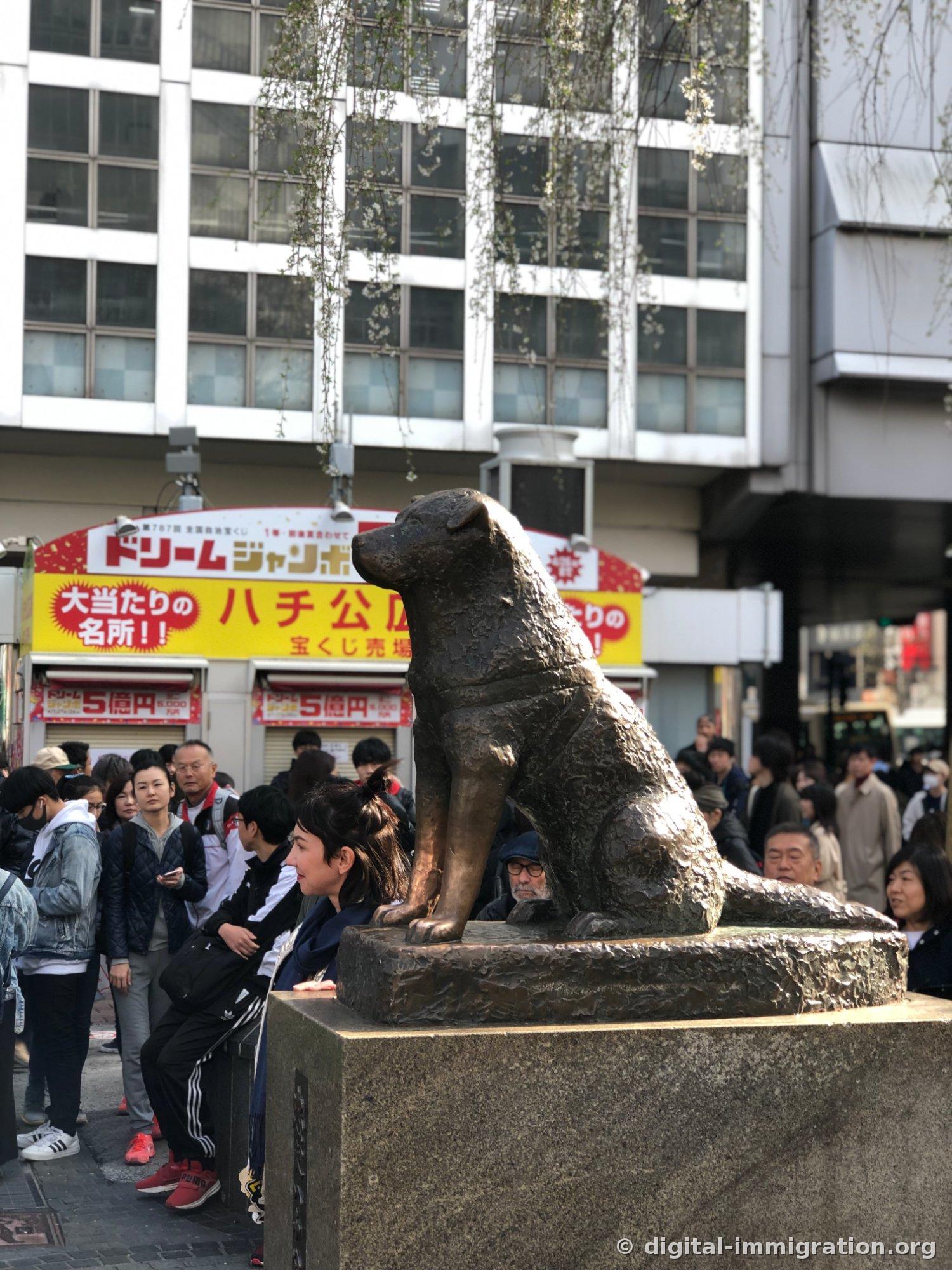 ハチ公像の横姿、後ろに「ハチ公」の黄色い宝くじ販売所の看板と人々が見える