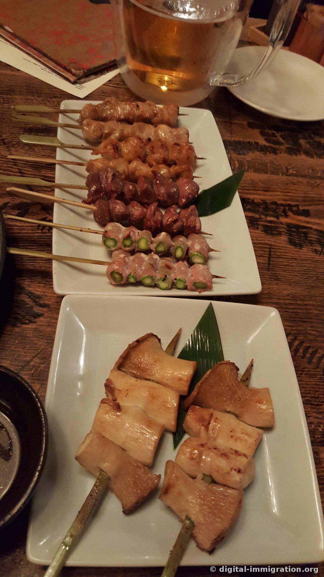 Assorted yakitori skewers and asparagus bacon wrap on a white square plate, with king trumpet mushroom bacon wrap skewers on a plate below, a draft beer glass in the background, all on a wooden table