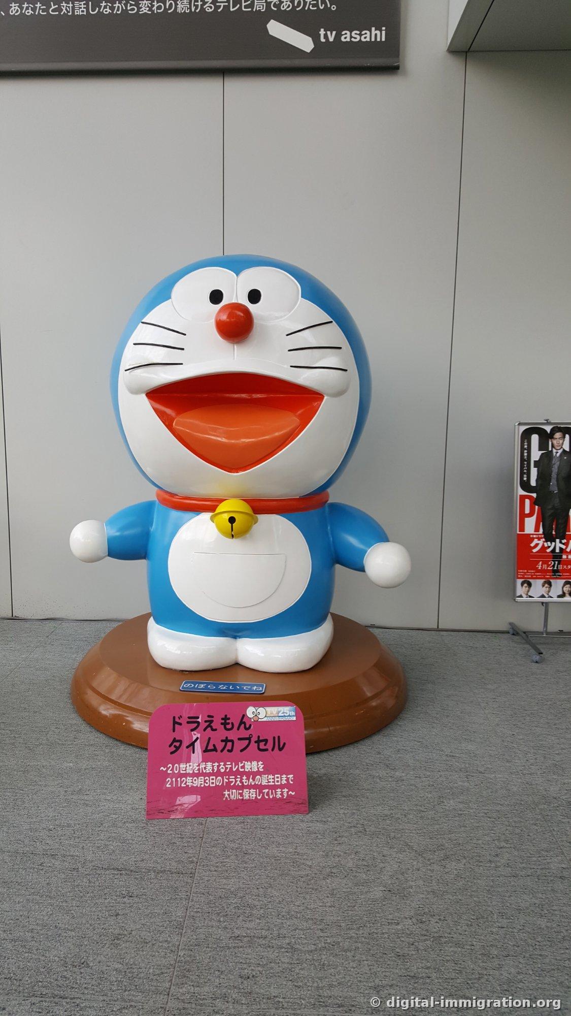Figurine de Doraemon se dressant sur un socle en bois devant un mur gris avec une pancarte rose 'Capsule temporelle de Doraemon' à ses pieds, et un panneau signalétique 'tv asahi' logo au-dessus