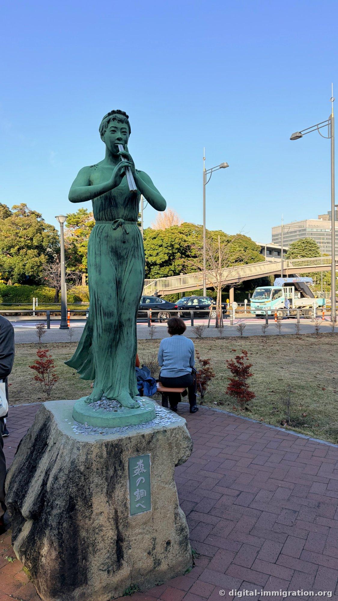 Une statue de femme en bronze jouant de la flûte avec beaucoup de pièces de monnaie accumulées au pied, derrière on voit la route et un passage surélevé