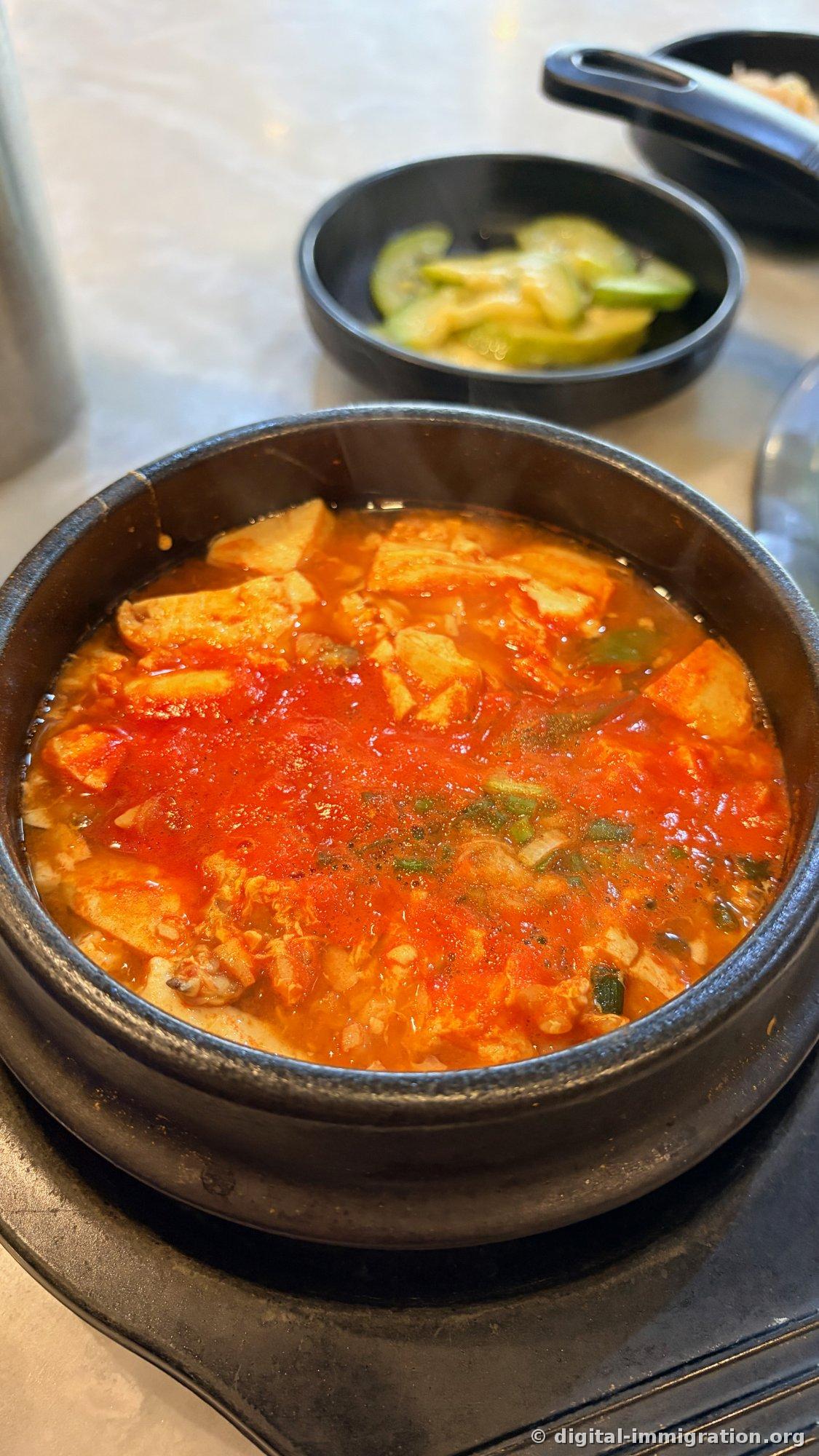 Sundubu-jjigae rouge en pleine ébullition dans un ttukbaegi, avec de gros morceaux de tofu, des coquillages et de la ciboule émincée. En arrière-plan, une assiette de concombre assaisonné.