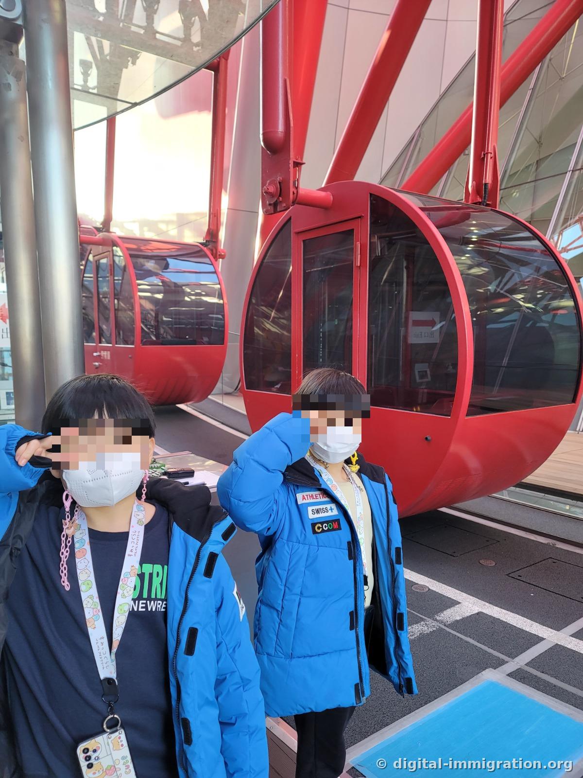 屋外乗降場で赤い観覧車カプセルの前に、青いダウンジャケットを着た子ども二人がVサインをして立っている様子