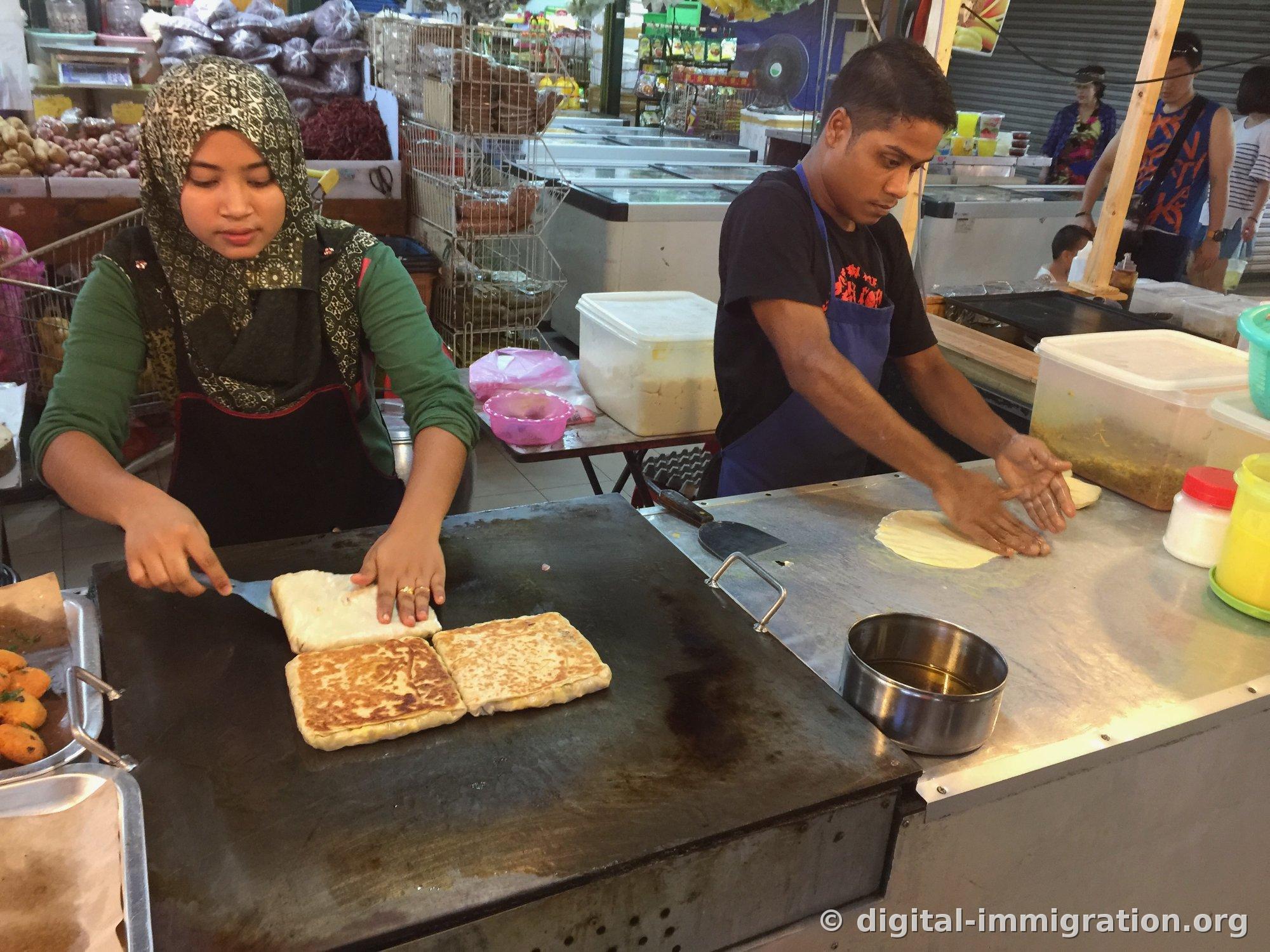 A roti canai stall with a woman folding roti with a spatula and a man stretching dough by hand at a griddle