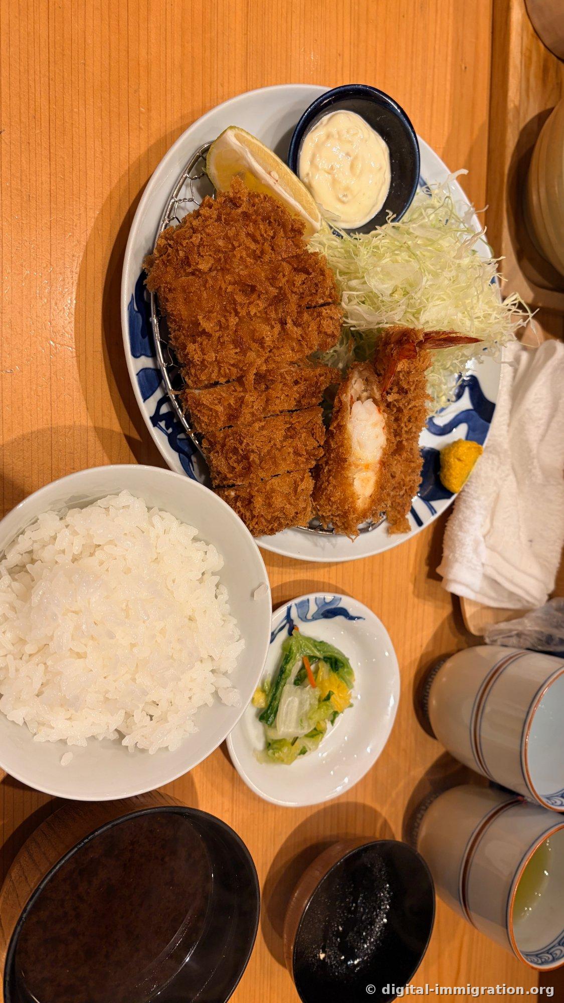 从上往下看的一套。猪排和虾炸块的盘子、堆得很高的白米饭、细切的青菜泡菜、两碗汤和绿茶杯