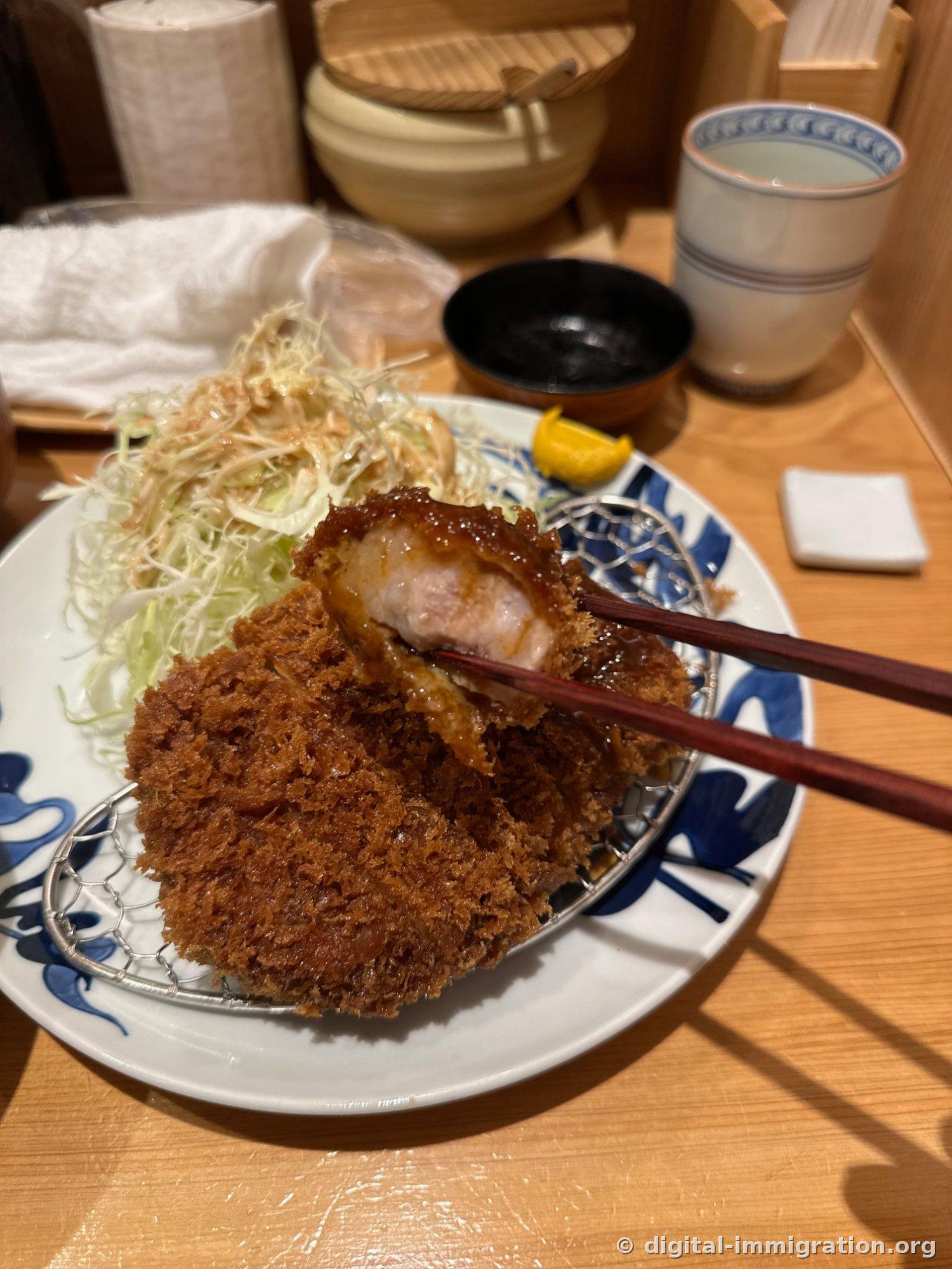 淋上炸猪排酱油的里脊肉炸猪排被筷子夹起，旁边是柚子块和淋过芝麻酱汁的卷心菜