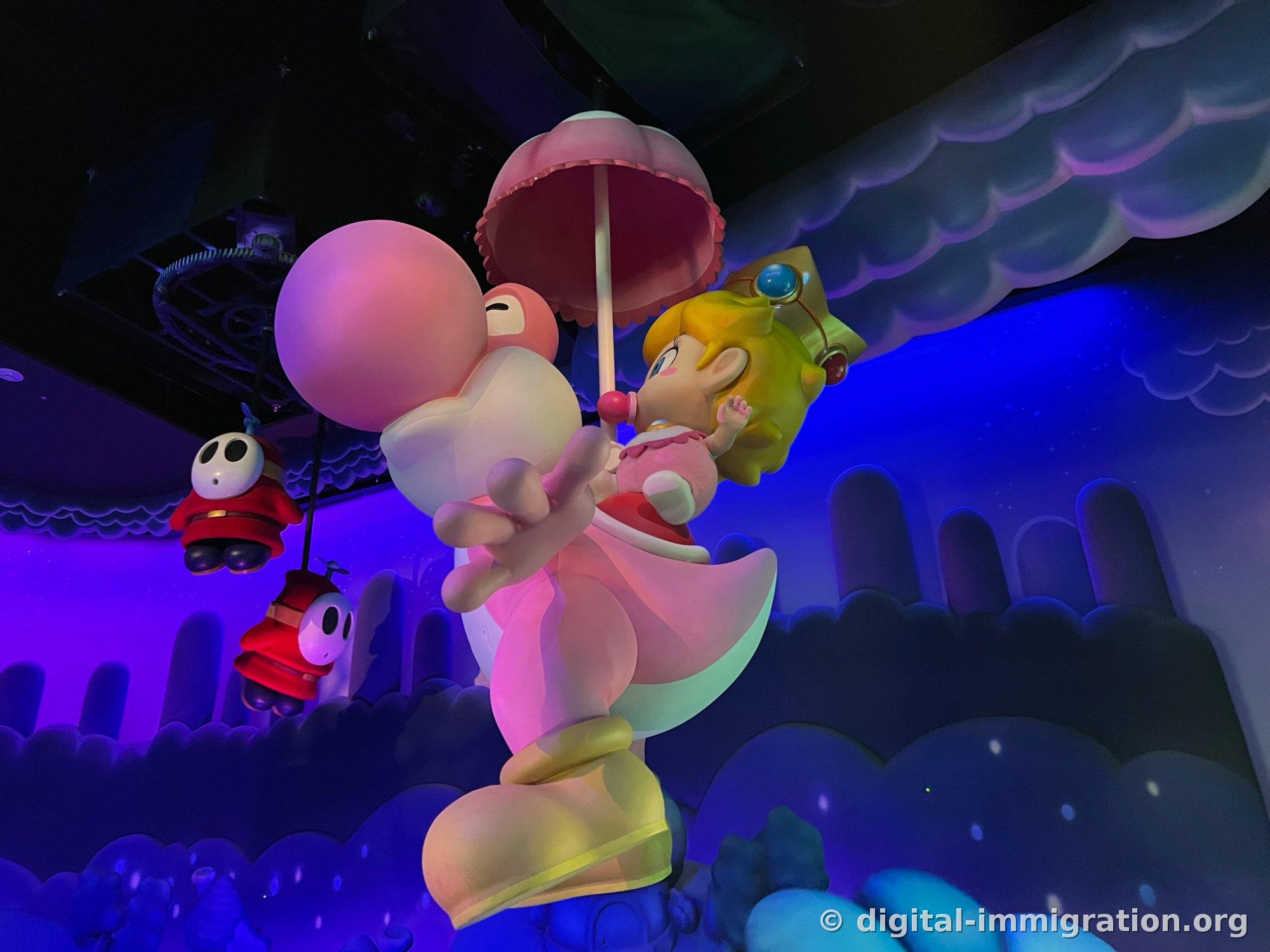 An indoor sculpture of a pink Yoshi holding a pink umbrella and cradling Baby Peach wearing a crown, floating above clouds